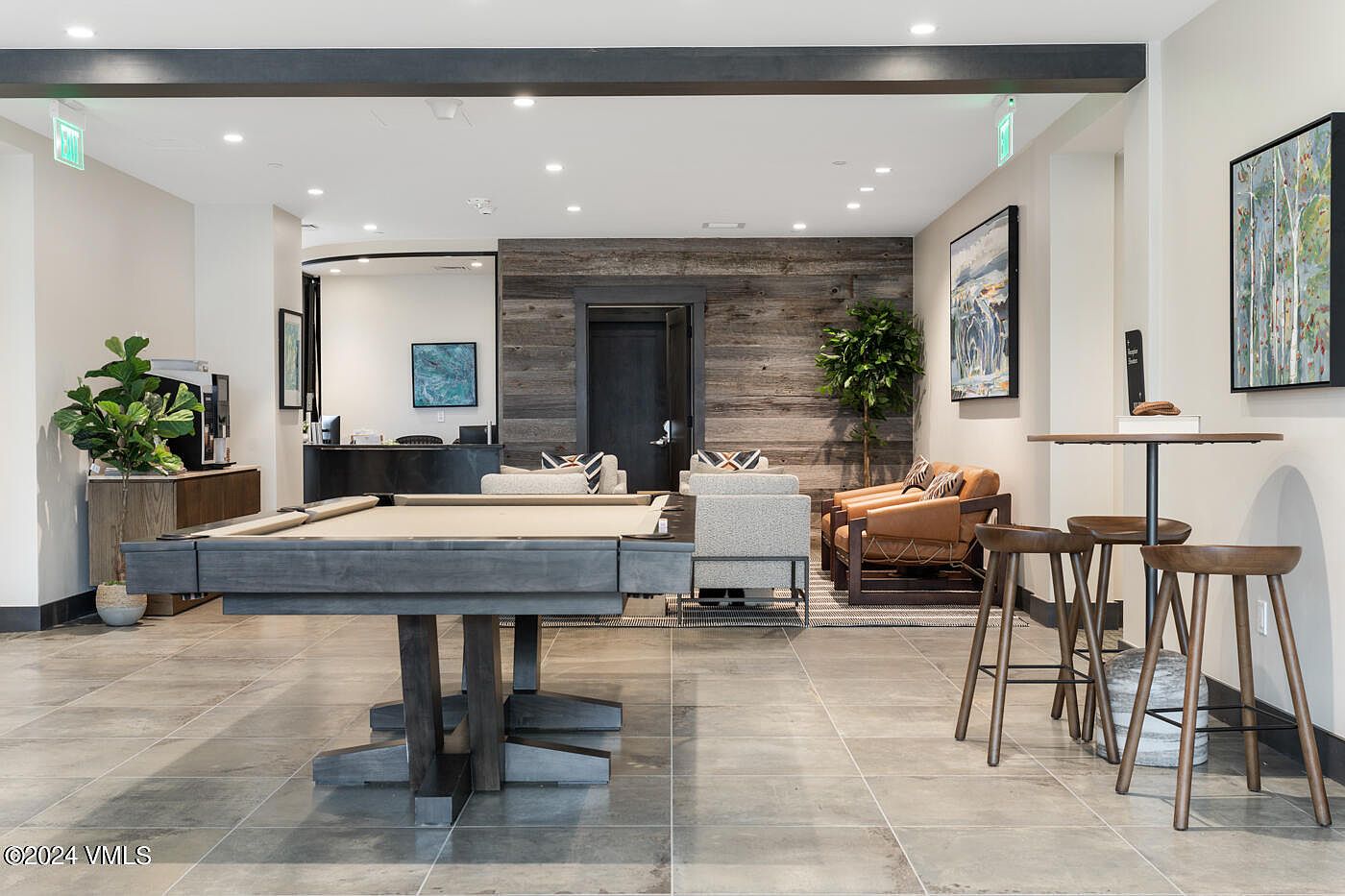 This inviting community lounge features a contemporary design with a prominent pool table in the foreground and a cozy seating area accented by a rustic wood-paneled wall. The space is furnished with modern armchairs and leather seating, creating a sophisticated yet relaxed atmosphere perfect for social gatherings. The open-concept layout is complemented by neutral tile flooring, recessed lighting, and tasteful wall art, offering a high-end amenity experience.