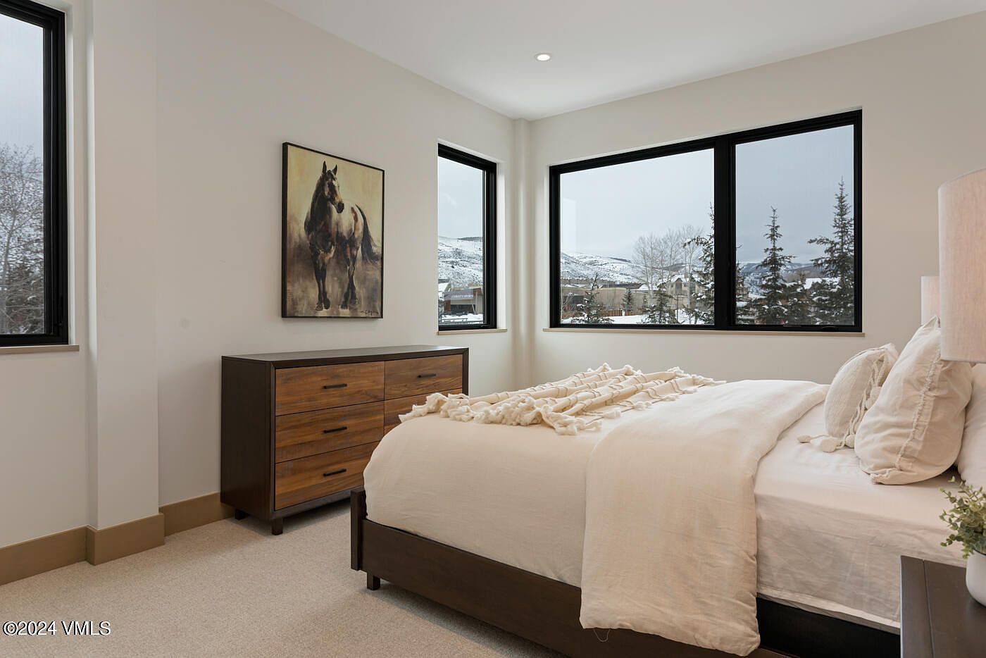 This bright and modern bedroom features a neutral color palette, a comfortable bed with a textured throw, and a dark wood dresser adorned with a horse-themed painting. Large windows frame a scenic, snow-covered mountain view, filling the room with natural light. The clean lines and minimalist decor create a serene and inviting atmosphere.