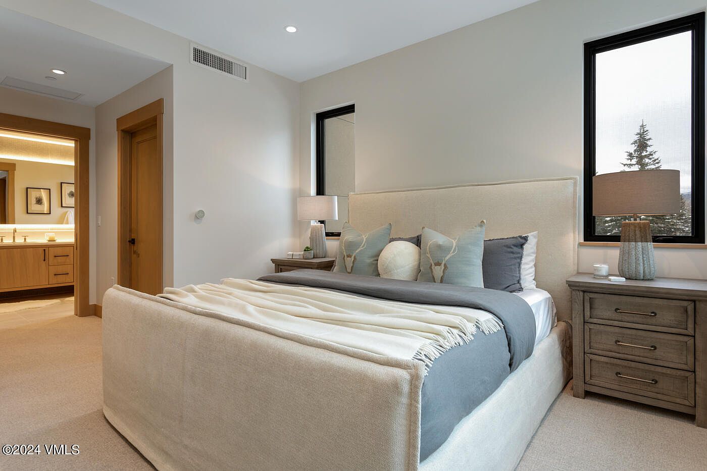 This elegant primary bedroom features a neutral color palette with a plush upholstered bed as the focal point, accented by decorative pillows and a soft throw blanket. The room is flanked by matching wooden nightstands with textured table lamps, while a large window provides natural light and a view of the snowy outdoors. The space feels serene and sophisticated, with a seamless transition into an adjacent bathroom visible through the doorway.