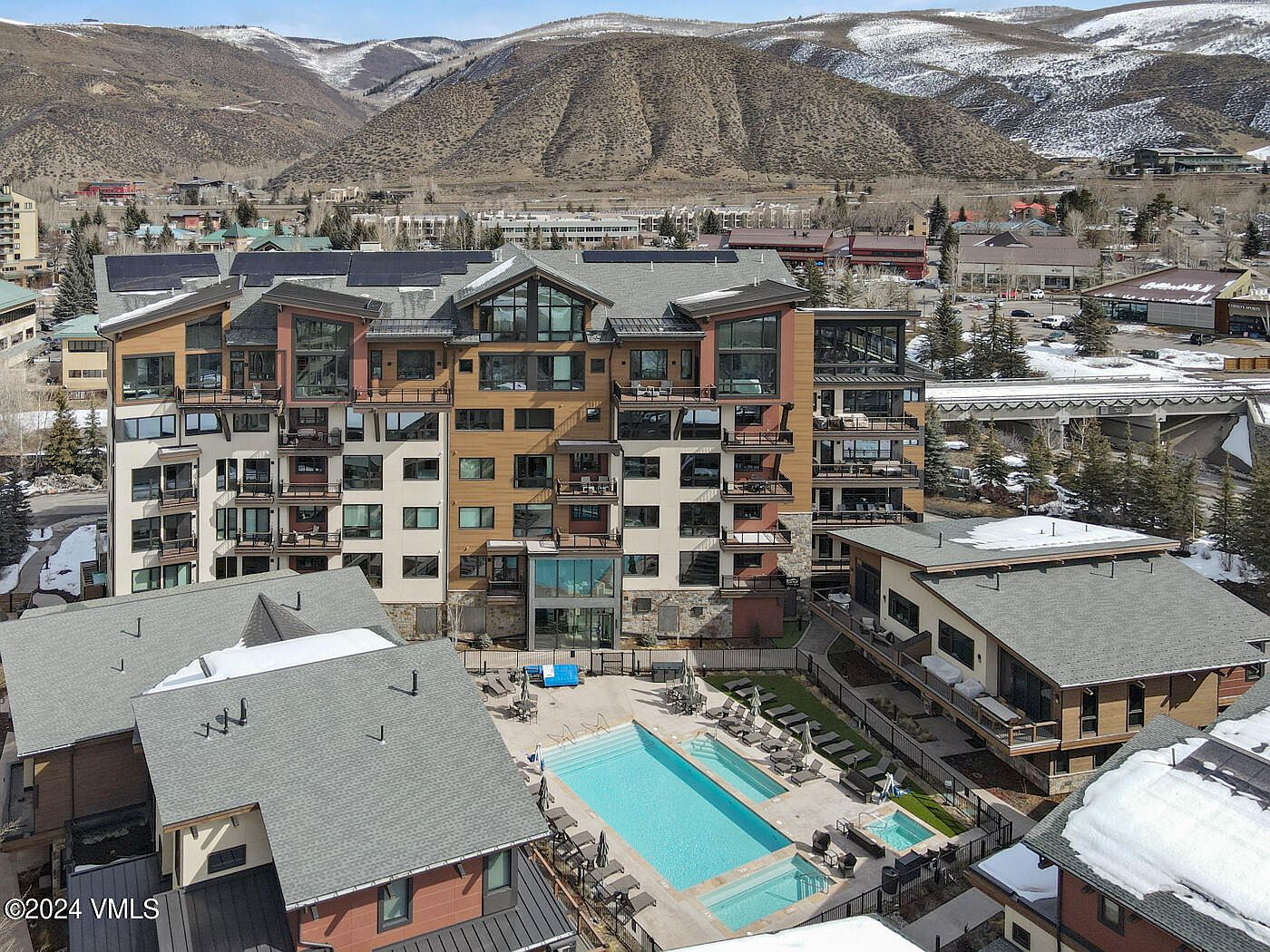 This high-angle aerial shot captures a multi-story mountain resort complex featuring a central swimming pool and hot tub area surrounded by modern, multi-level residential buildings. The architecture blends contemporary design with rustic mountain aesthetics, set against a backdrop of snow-dusted hills and a clear blue sky. The scene conveys a luxurious, year-round vacation atmosphere with well-maintained outdoor amenities.