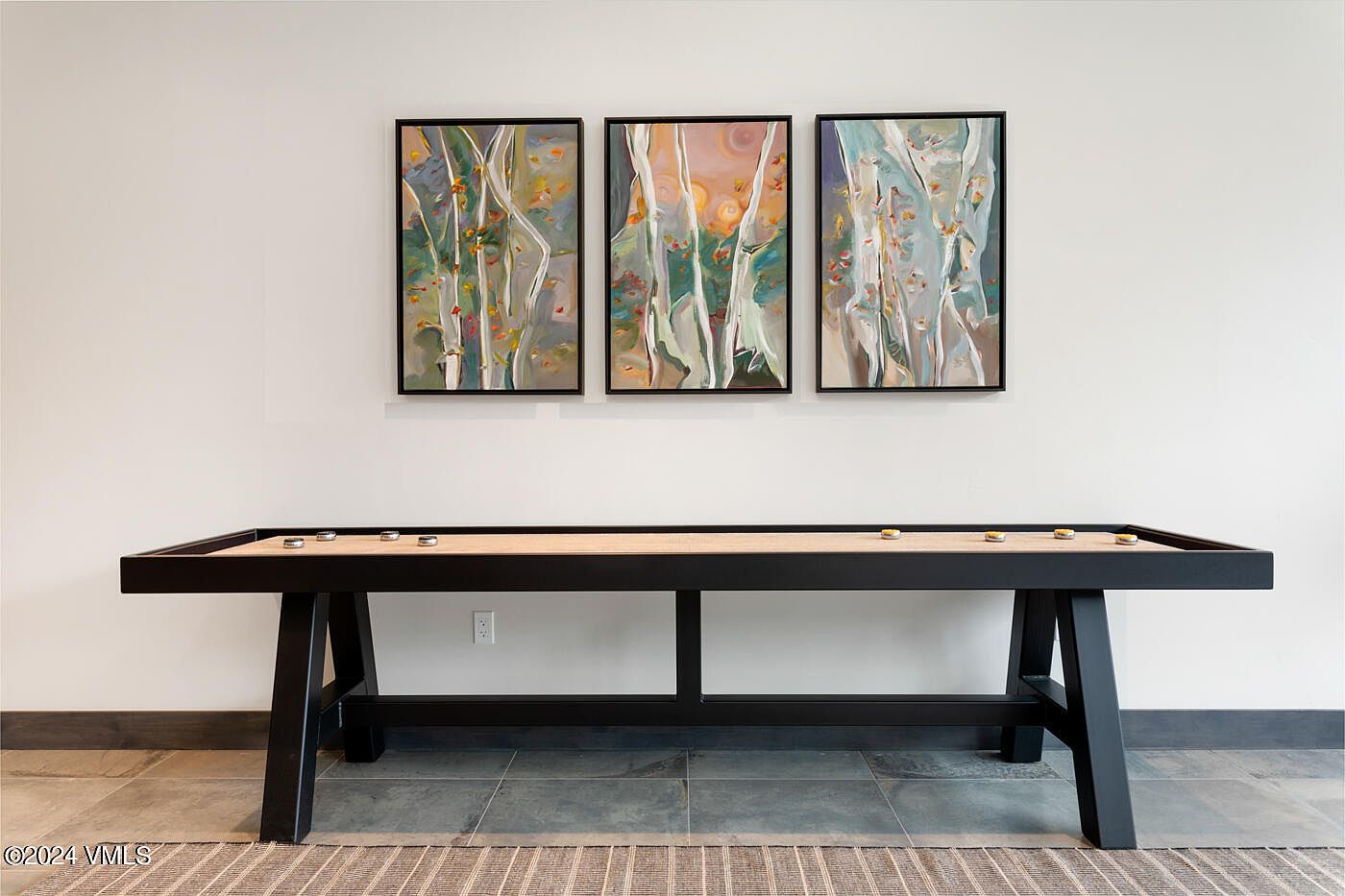 This image features a modern shuffleboard table centered against a minimalist white wall, adorned with a triptych of abstract birch tree paintings. The scene is captured from a straight-on, eye-level perspective, emphasizing the clean lines of the black-framed table and the artistic focal point above it. The neutral color palette and simple composition create a sophisticated, recreational atmosphere suitable for a game room or lounge area.