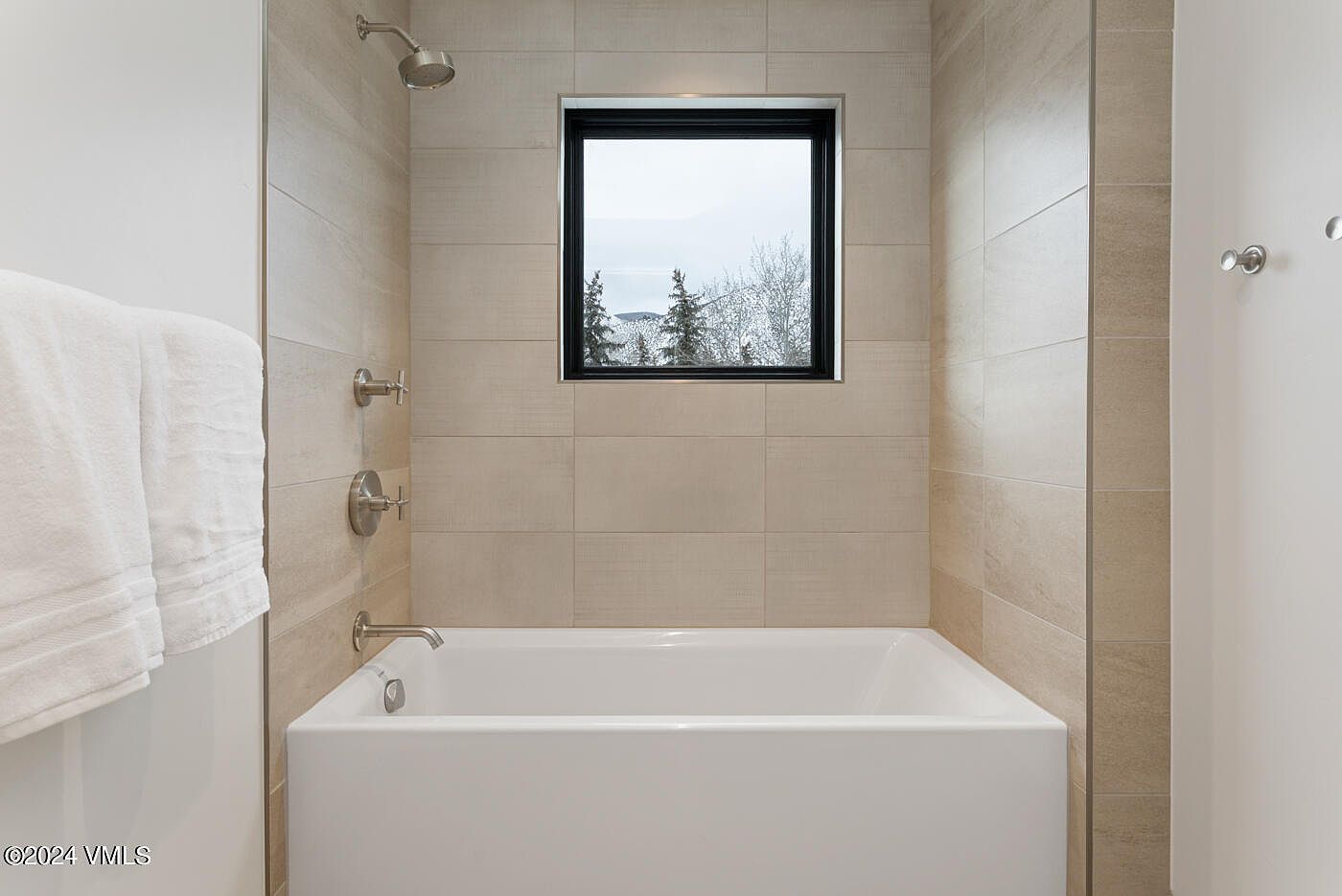 This modern bathroom features a clean, minimalist design with light-toned, large-format wall tiles surrounding a deep white soaking tub. A central window provides natural light and a scenic view of snow-covered trees, while polished chrome fixtures add a sleek, contemporary touch. The space feels bright, serene, and well-maintained, perfect for a relaxing spa-like experience.