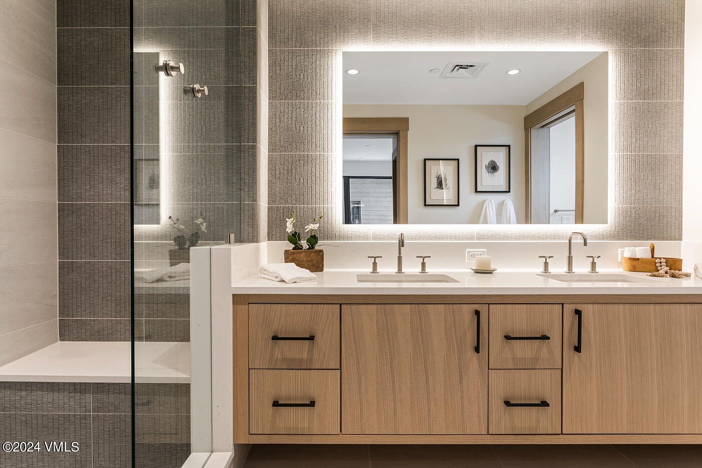 This modern primary bathroom features a sleek double vanity with light wood cabinetry, white quartz countertops, and a large, illuminated mirror. To the left, a glass-enclosed walk-in shower with textured grey tile walls adds a sophisticated, spa-like aesthetic. The clean lines and neutral color palette create a bright, contemporary, and luxurious atmosphere.