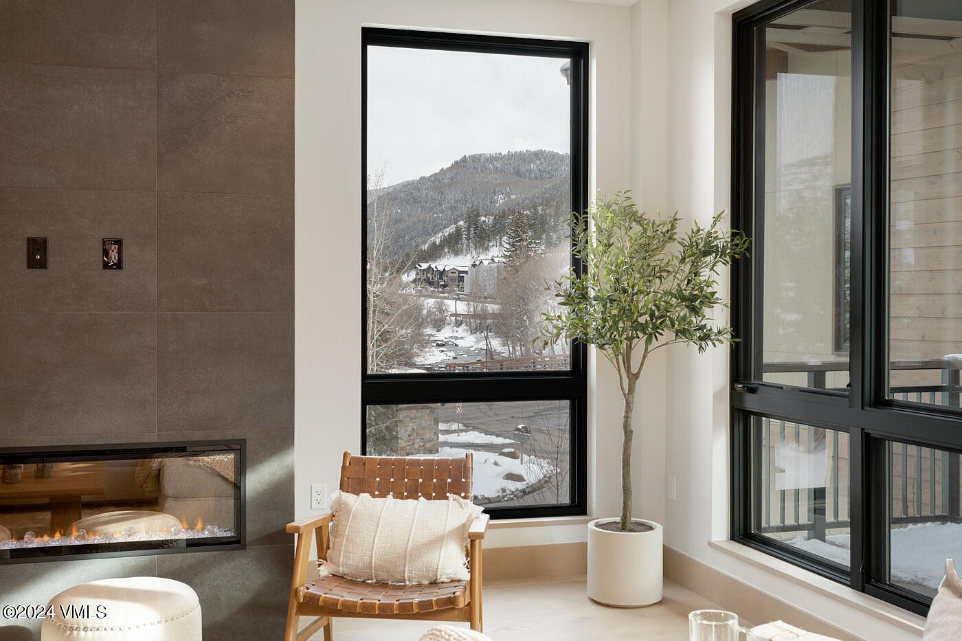 This modern living room features a sleek, floor-to-ceiling gray tiled fireplace and a comfortable leather-accented chair, creating a cozy yet sophisticated atmosphere. A large window provides a stunning, panoramic view of a snow-covered mountain landscape, while a potted olive tree adds a touch of natural greenery to the bright, minimalist space. The composition emphasizes the seamless blend of contemporary interior design with the breathtaking natural scenery outside.