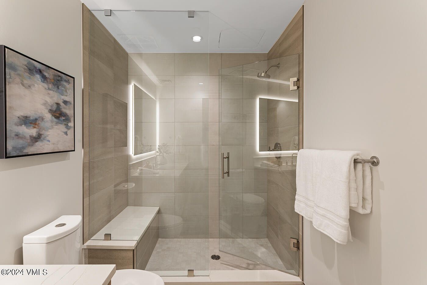 This modern, spa-inspired bathroom features a spacious walk-in shower with large-format beige tiles, a built-in bench, and a frameless glass enclosure. The space is accented by sleek, illuminated mirrors and high-end fixtures, creating a clean and sophisticated aesthetic. A white towel hangs conveniently on a rack, emphasizing the functional yet luxurious design of the room.