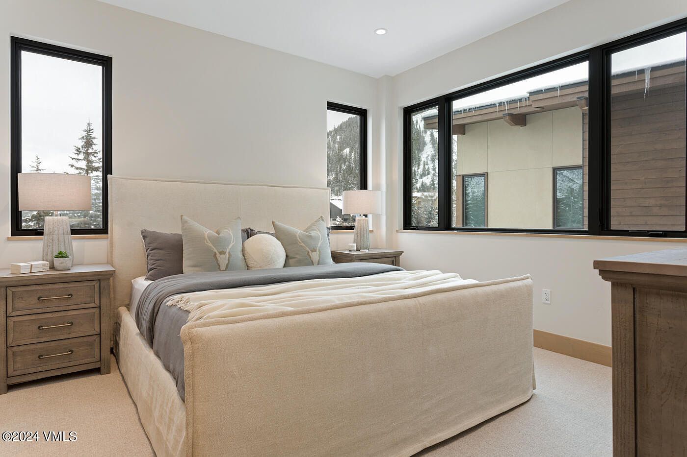 This serene primary bedroom features a neutral color palette with a plush upholstered bed as the focal point, accented by soft grey and cream bedding. Large, black-framed windows flood the room with natural light and offer scenic views of a snowy, mountainous landscape. The space is completed by matching wooden nightstands and lamps, creating a warm and inviting atmosphere.
