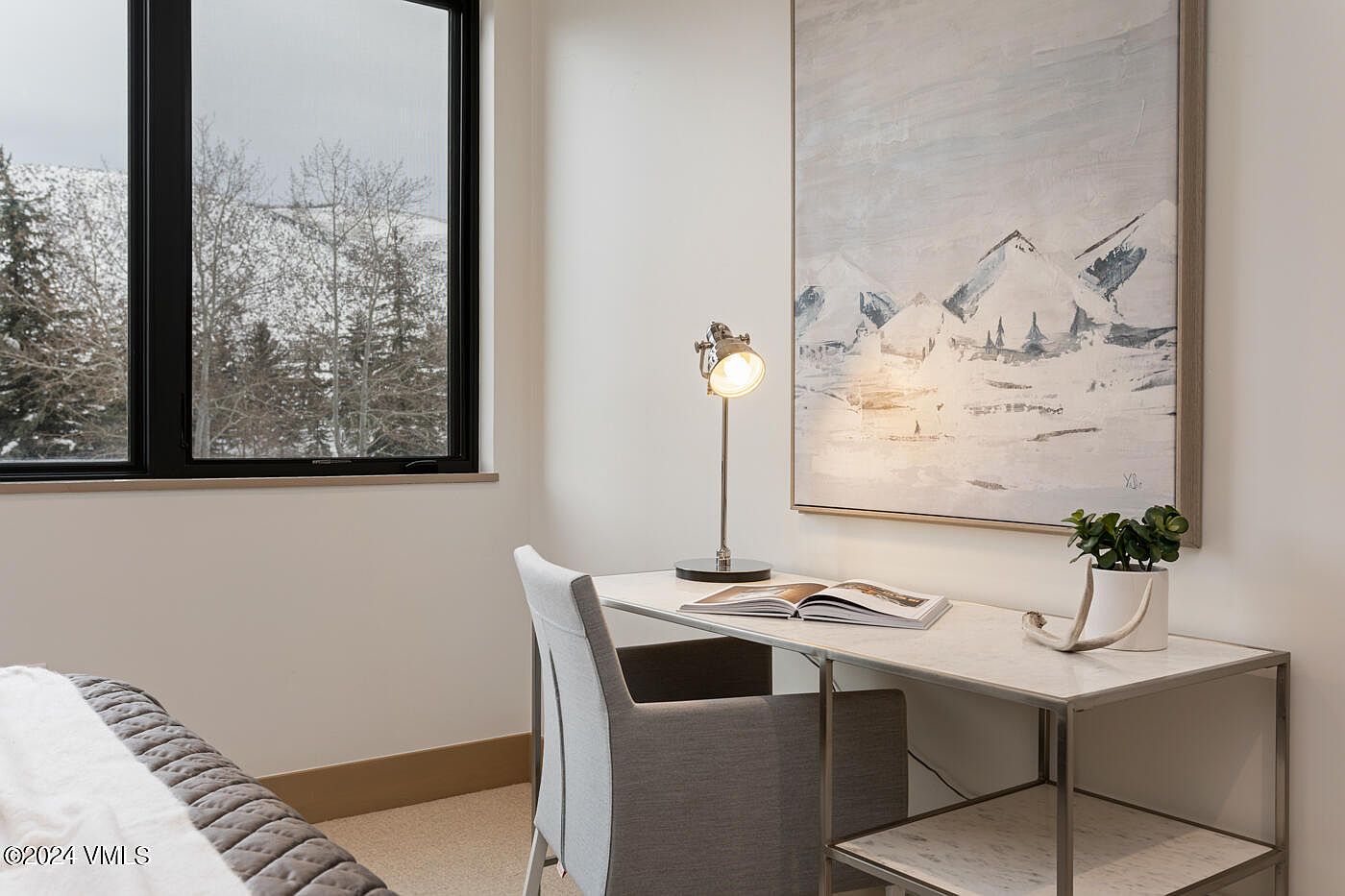 This bright and serene home office features a sleek, marble-topped desk paired with a modern gray upholstered chair. A large, abstract mountain-themed painting hangs on the wall, complementing the snowy mountain view visible through the adjacent window. The space is accented by a minimalist desk lamp and a small potted plant, creating a calm and productive atmosphere.