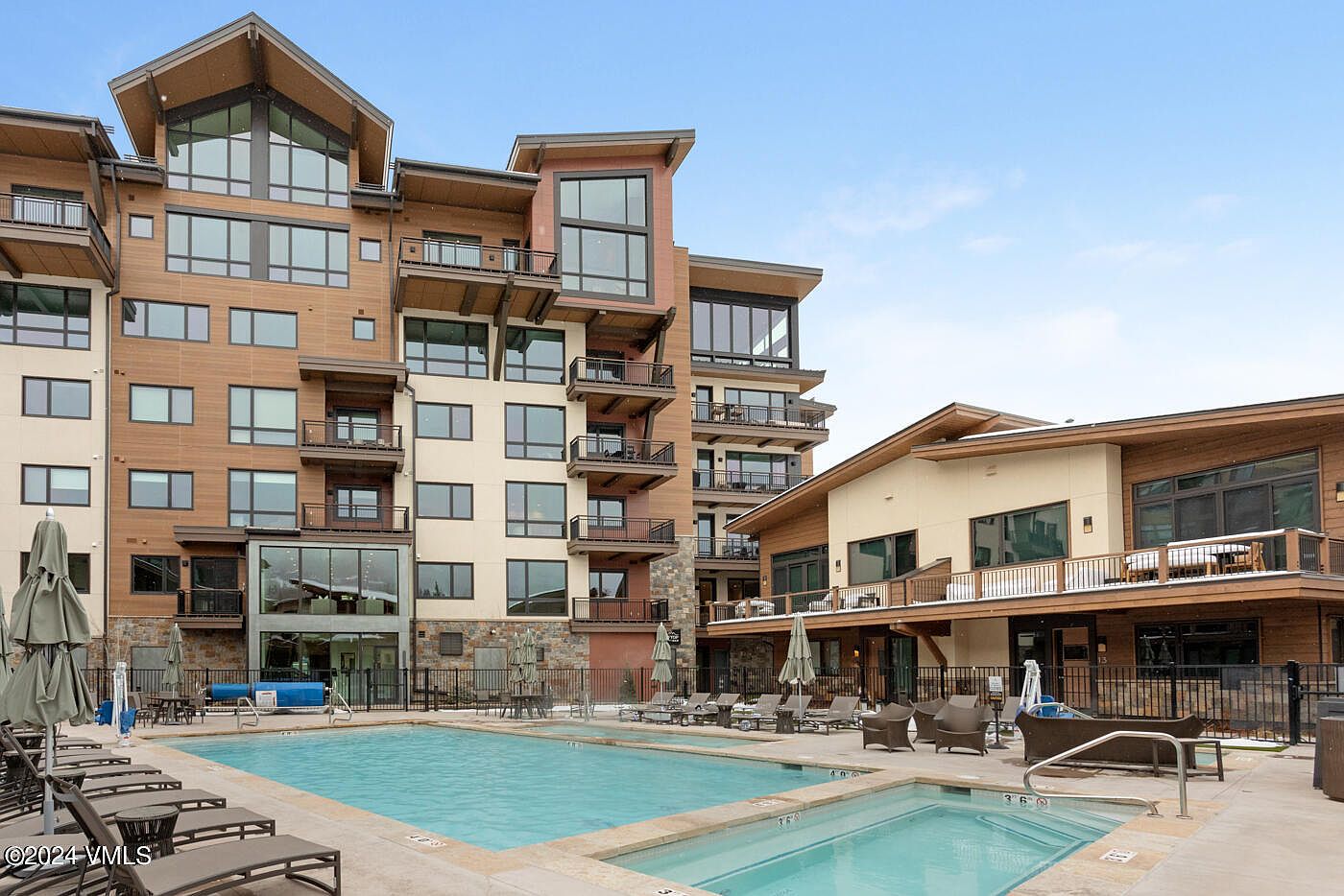 This image captures a luxurious outdoor pool and spa area set against a multi-story, modern mountain-style resort building. The scene features a clear blue swimming pool in the foreground, complemented by a smaller hot tub, surrounded by lounge chairs and patio furniture. The architecture showcases a blend of wood siding, stone accents, and large glass windows, creating an inviting and upscale atmosphere for residents or guests.