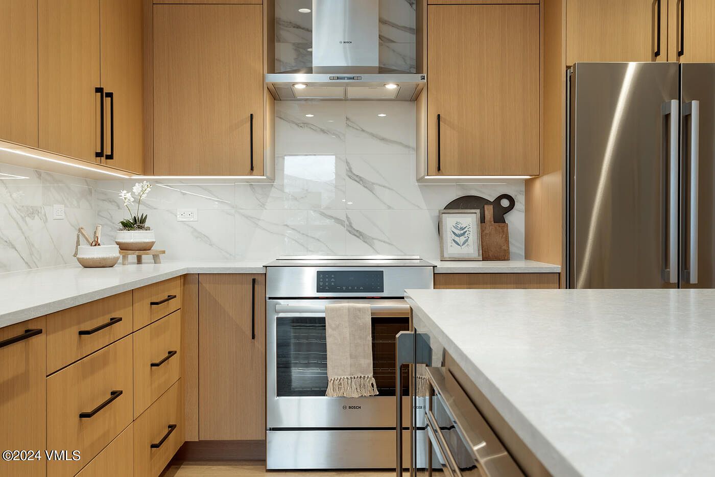 This modern kitchen features warm wood-toned cabinetry paired with sleek, light-colored quartz countertops and a striking marble-patterned backsplash. The space is equipped with high-end stainless steel Bosch appliances, including a range and a range hood, creating a clean and professional aesthetic. The perspective is a straight-on eye-level shot that highlights the functional layout and the sophisticated, contemporary design of the cooking area.