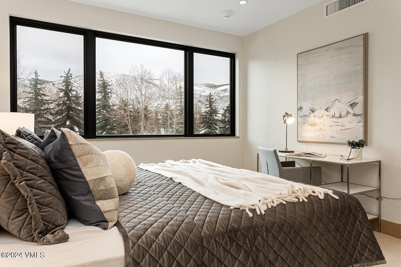 This cozy bedroom features a plush, quilted dark grey bedspread and a variety of textured pillows, creating a warm and inviting atmosphere. A sleek desk area is positioned near a large window that offers a stunning, panoramic view of a snow-covered mountain landscape. The room is accented by a large, abstract mountain-themed painting and minimalist decor, emphasizing a modern, serene mountain retreat aesthetic.