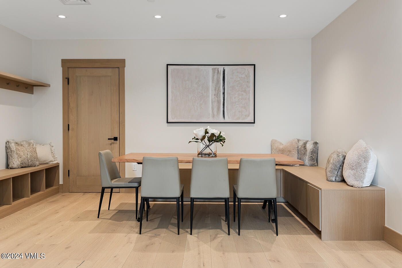This contemporary dining area features a natural wood table paired with four sleek, light grey upholstered chairs and a built-in corner bench with plush, textured pillows. The minimalist aesthetic is complemented by light hardwood flooring, a neutral color palette, and a large abstract piece of wall art. The scene is captured from a straight-on, eye-level perspective, emphasizing the clean lines and functional, modern design of the space.