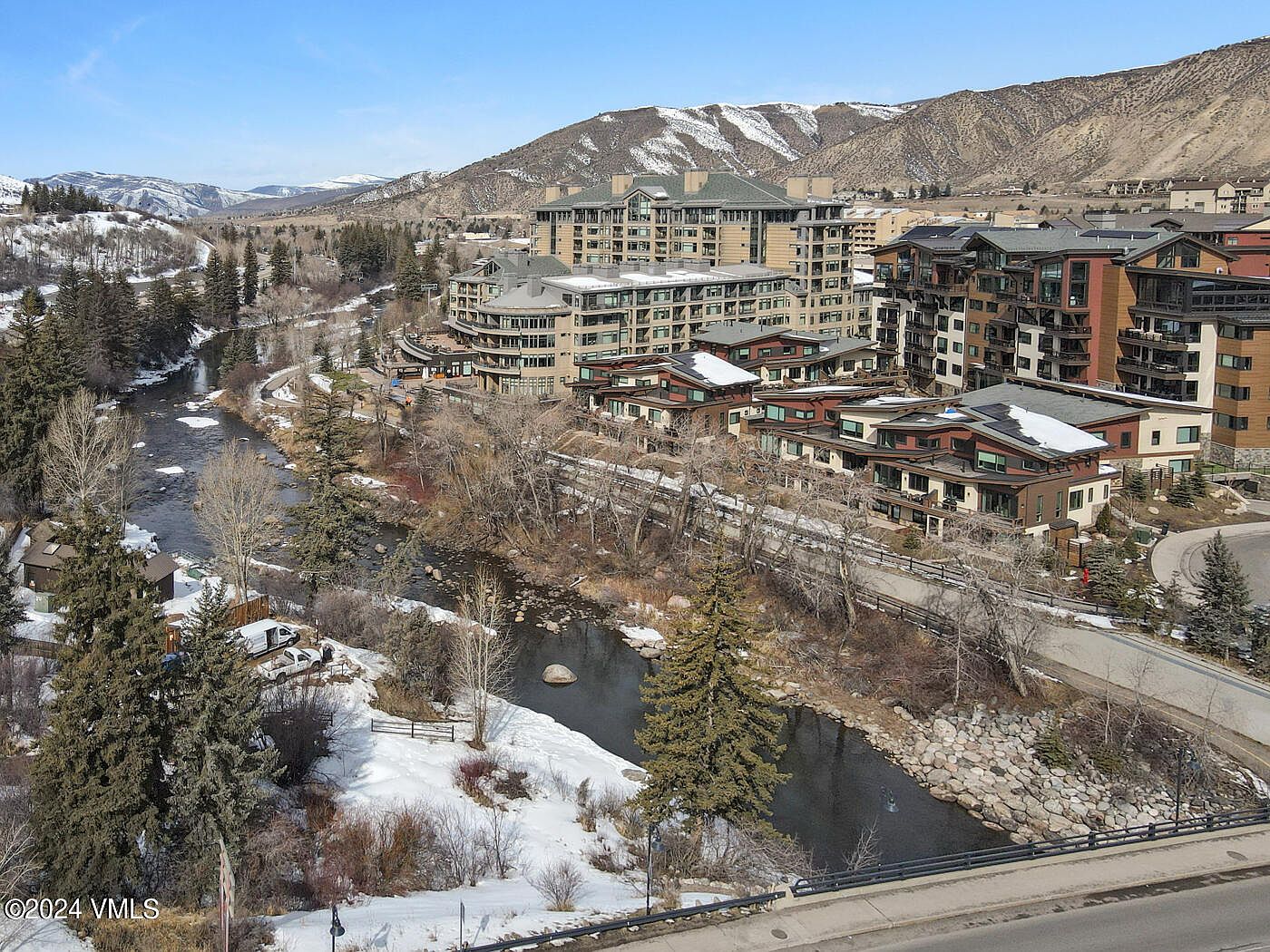 This high-angle aerial view captures a scenic mountain resort community nestled along a winding river. The landscape features a mix of modern multi-story residential buildings, snow-dusted terrain, and evergreen trees, set against a backdrop of rugged, snow-capped mountains. The perspective emphasizes the integration of luxury living with the natural environment, highlighting the proximity to both the water and the surrounding wilderness.