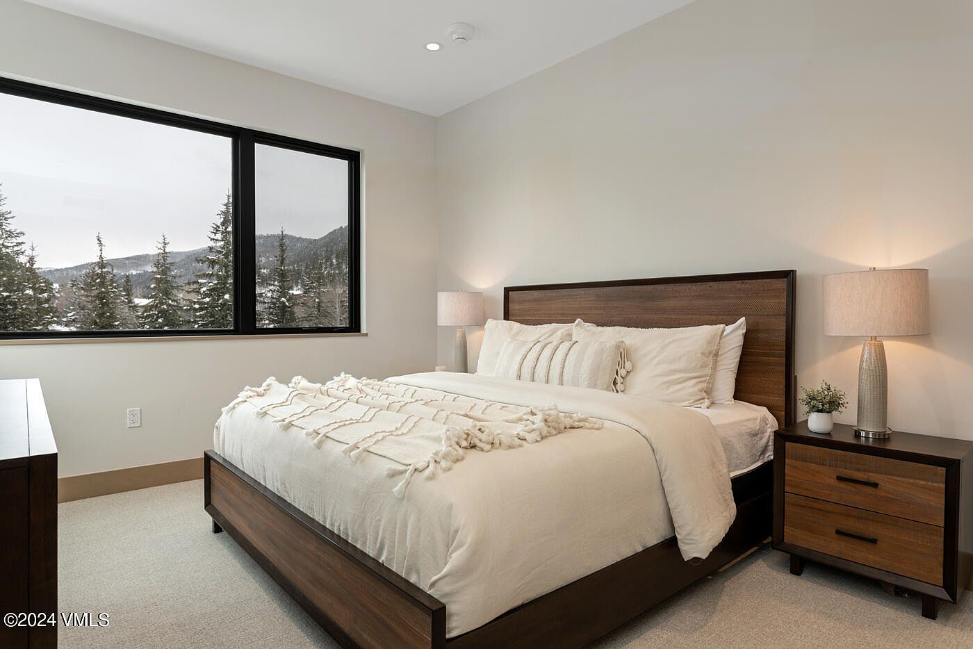 This modern primary bedroom features a sleek wooden bed frame with a plush, neutral-toned bedding set and a textured throw blanket. A large window provides a stunning, panoramic view of a snow-covered mountain landscape, while warm bedside lamps create a cozy and inviting atmosphere. The room is finished with clean, minimalist decor and a dark wood nightstand, emphasizing a contemporary and serene aesthetic.