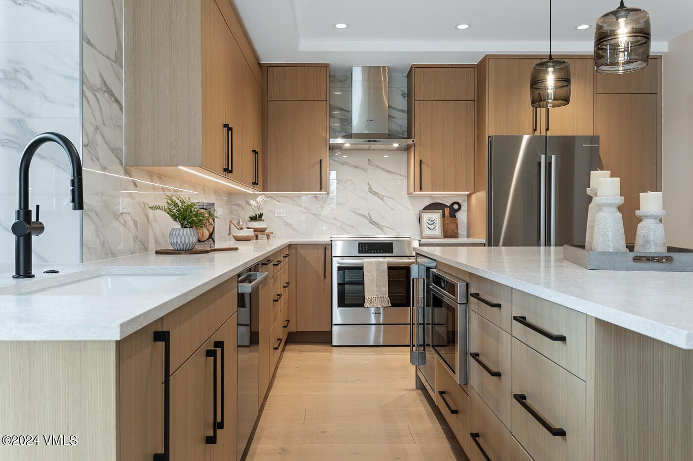 This modern, high-end kitchen features sleek light wood cabinetry, white marble-veined countertops, and premium stainless steel appliances. The space is illuminated by stylish pendant lights over the island and under-cabinet lighting, creating a warm and inviting atmosphere. The perspective is a straight-on, eye-level shot that highlights the efficient galley layout and contemporary design aesthetic.