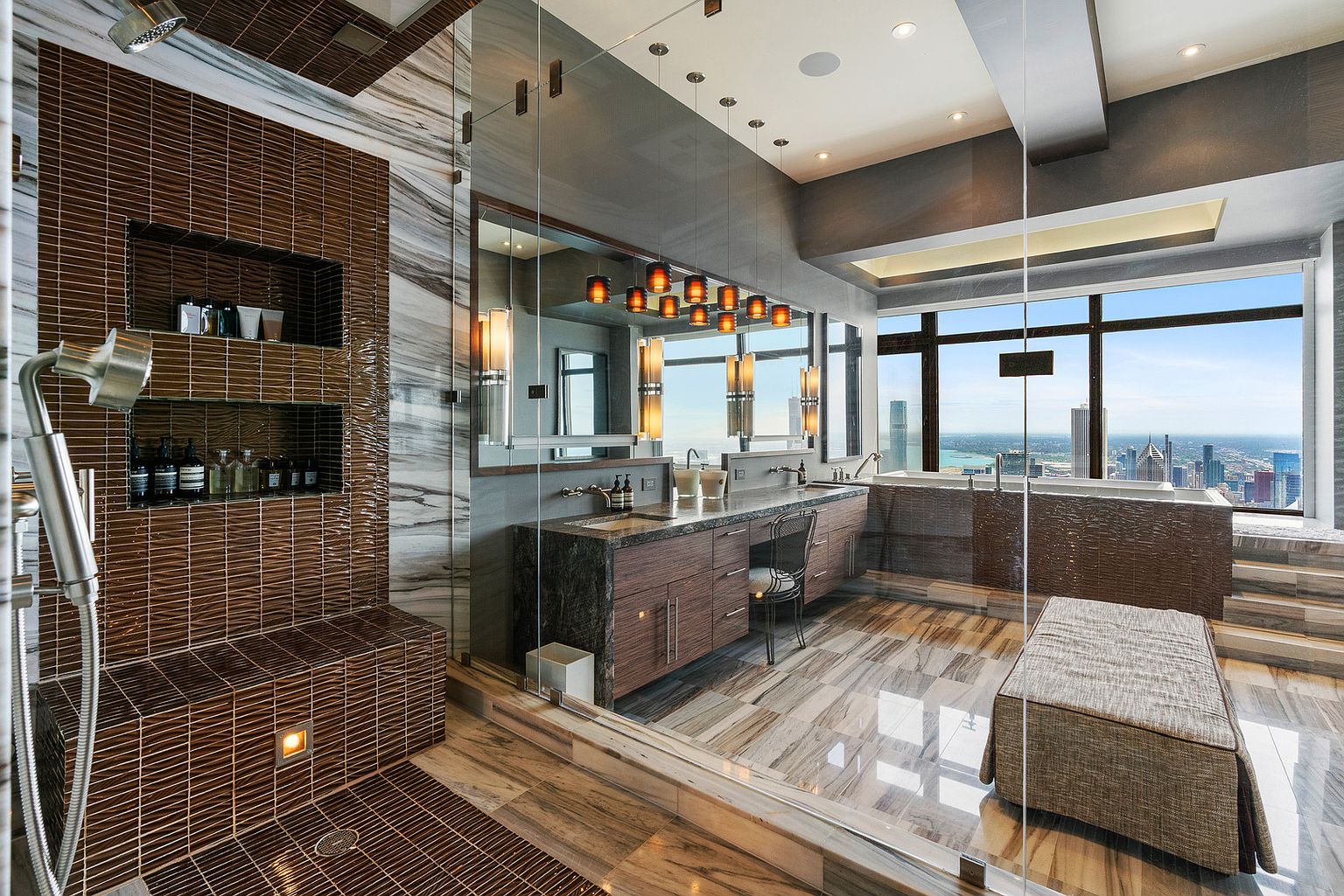 This luxurious primary bathroom features a glass-enclosed shower area with dark, textured tile walls and built-in shelving. A long vanity with a stone countertop and integrated sinks sits beneath a large mirror, complemented by a series of pendant lights. The space is defined by floor-to-ceiling windows that offer a panoramic view of the city skyline and a deep soaking tub positioned to take advantage of the vista.