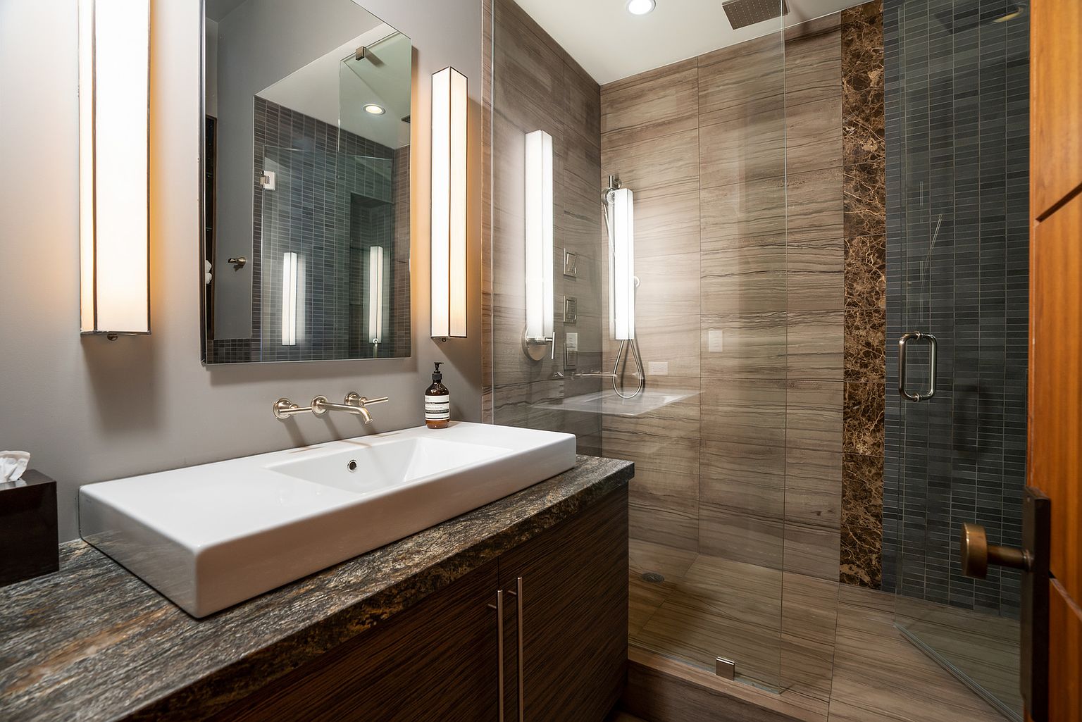 This modern bathroom features a sleek, rectangular vessel sink set atop a dark, textured vanity with a stone countertop. A large, frameless glass shower enclosure showcases horizontal wood-look tile walls accented by a vertical strip of decorative mosaic tile. The space is illuminated by two tall, vertical wall sconces flanking a rectangular mirror, creating a sophisticated and polished atmosphere.