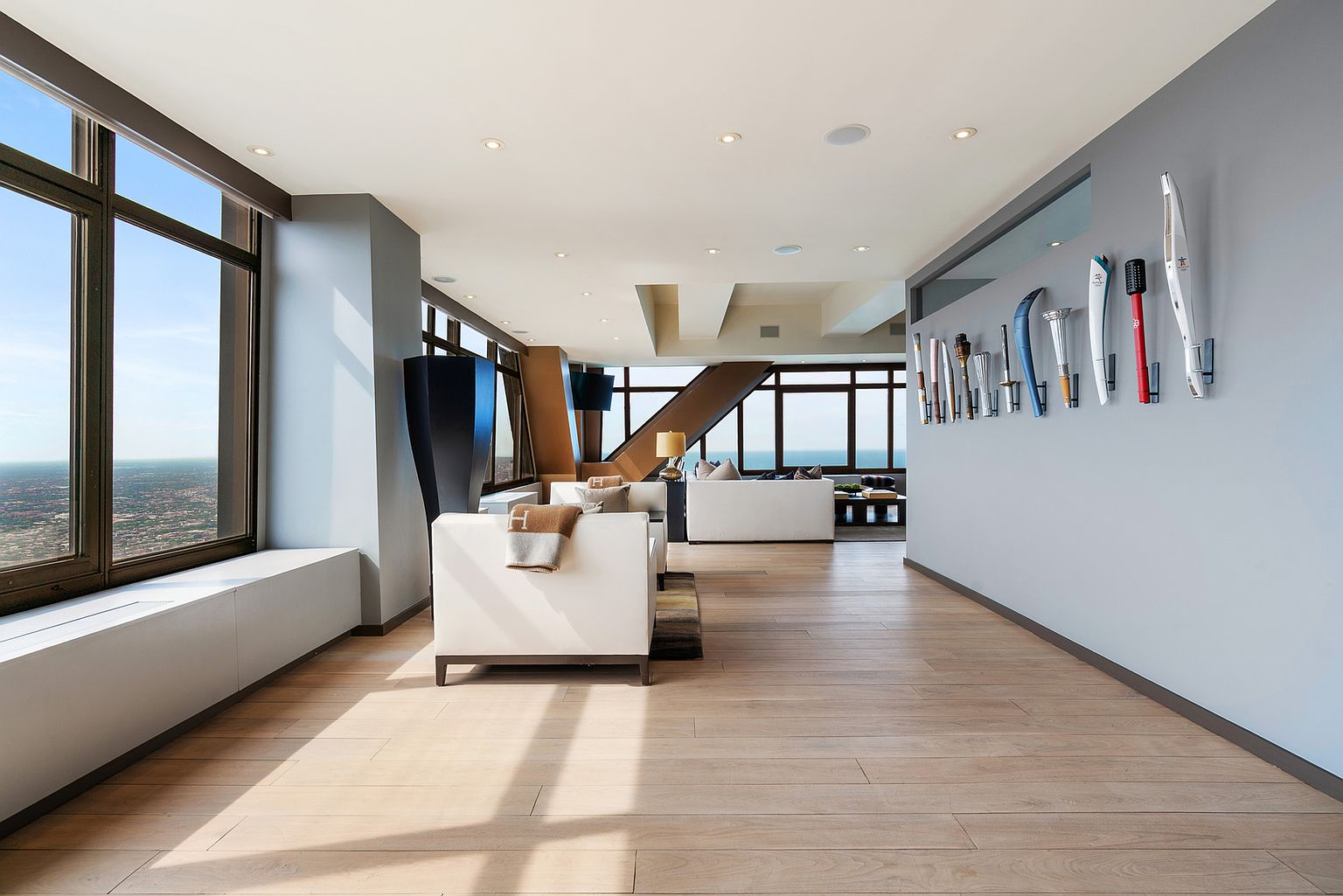 This expansive, high-rise living room features light wood flooring and a minimalist aesthetic, anchored by white sofas and a unique collection of Olympic torches displayed on a feature wall. Large windows provide panoramic city views, bathing the open-concept space in natural light. The room is designed with a sophisticated, modern layout that emphasizes clean lines and an airy, elevated atmosphere.