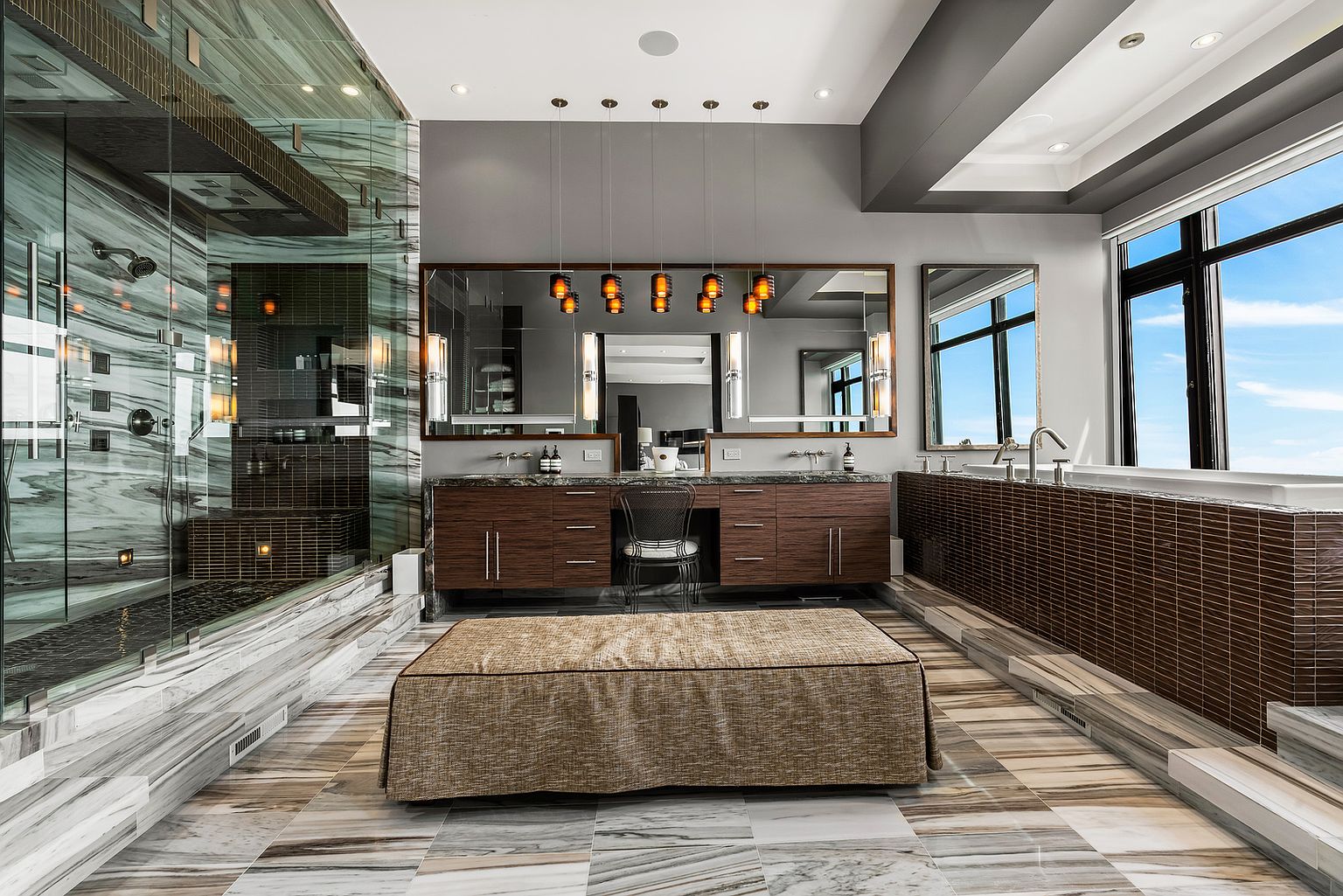 This luxurious primary bathroom features a large walk-in shower with patterned stone walls and a dark-tiled floor, positioned opposite a deep soaking tub set against a wall of dark, horizontal tiles. A central vanity with a dark wood finish and a makeup station sits beneath a wide mirror illuminated by five hanging pendant lights. A large, upholstered ottoman rests in the center of the room, which is finished with light-toned, veined stone flooring and large windows that provide ample natural light.