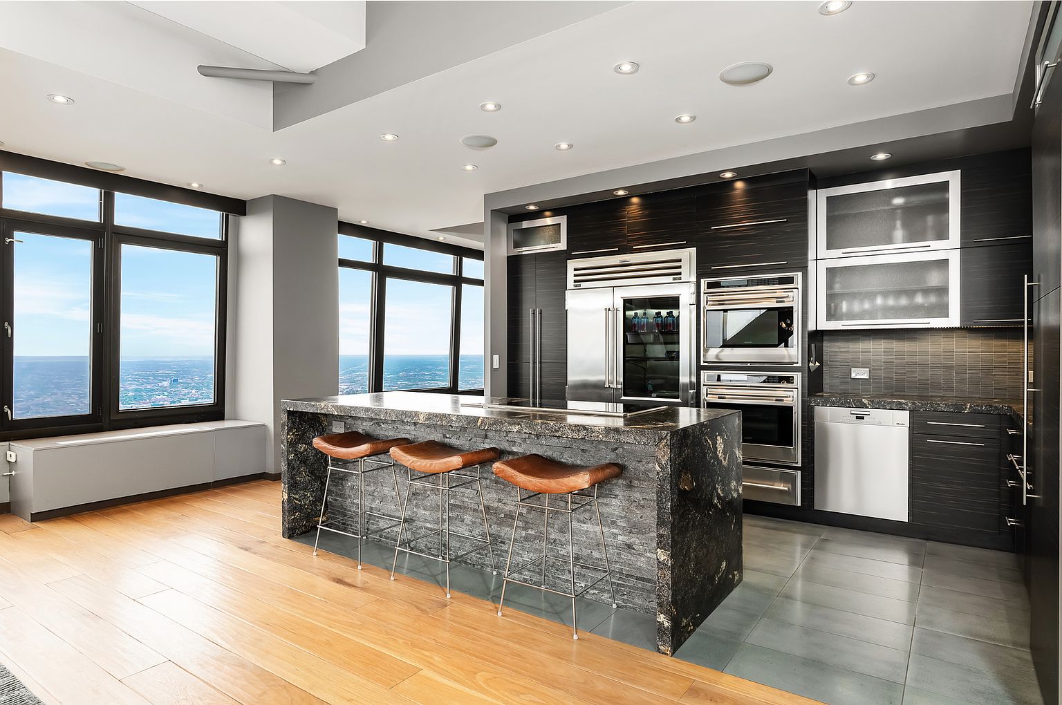 This sleek, modern kitchen features dark cabinetry, stainless steel appliances, and a large island with a stone countertop and three bar stools. The space is defined by a transition from light wood flooring to grey tile, with expansive windows providing a panoramic view of the city skyline. The overall aesthetic is sophisticated and high-end, emphasizing clean lines and a contemporary urban design.