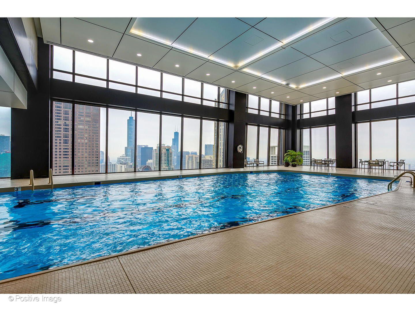 This indoor swimming pool area features expansive floor-to-ceiling windows that offer a panoramic view of the city skyline. The space is characterized by a neutral tiled deck, a large blue pool, and a modern ceiling design with recessed lighting. The perspective is wide and inviting, highlighting the luxury amenity and the impressive urban backdrop.