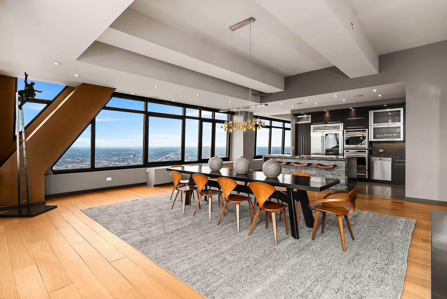 This expansive, open-concept dining area features a long, dark wood table surrounded by mid-century modern style chairs, set atop a large textured area rug. The space is defined by a wall of large windows that provide a panoramic view of the city skyline, complemented by a modern linear chandelier hanging above the table. In the background, a sleek kitchen with dark cabinetry and a stone-topped island creates a sophisticated, high-rise living atmosphere.