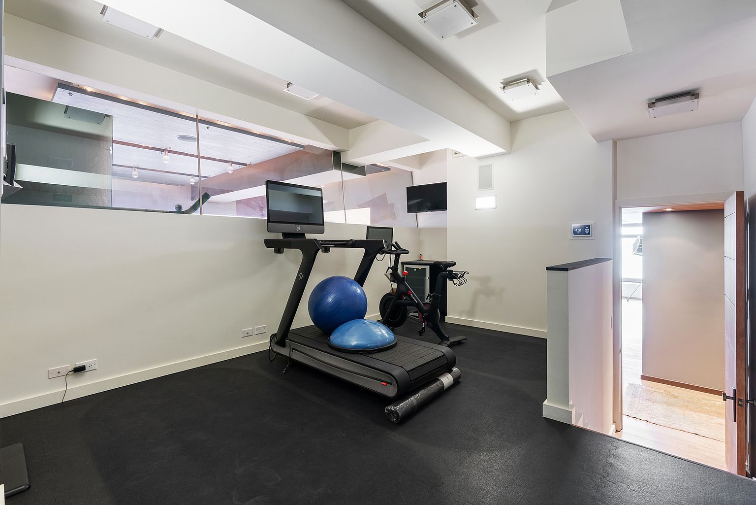 This home gym features a dedicated workout space with black rubber flooring, equipped with a treadmill and a stationary bike. The room includes a blue exercise ball and a balance trainer, with a glass partition overlooking an adjacent area. A doorway leads to a hallway with light-toned flooring, and the space is illuminated by recessed ceiling lights.
