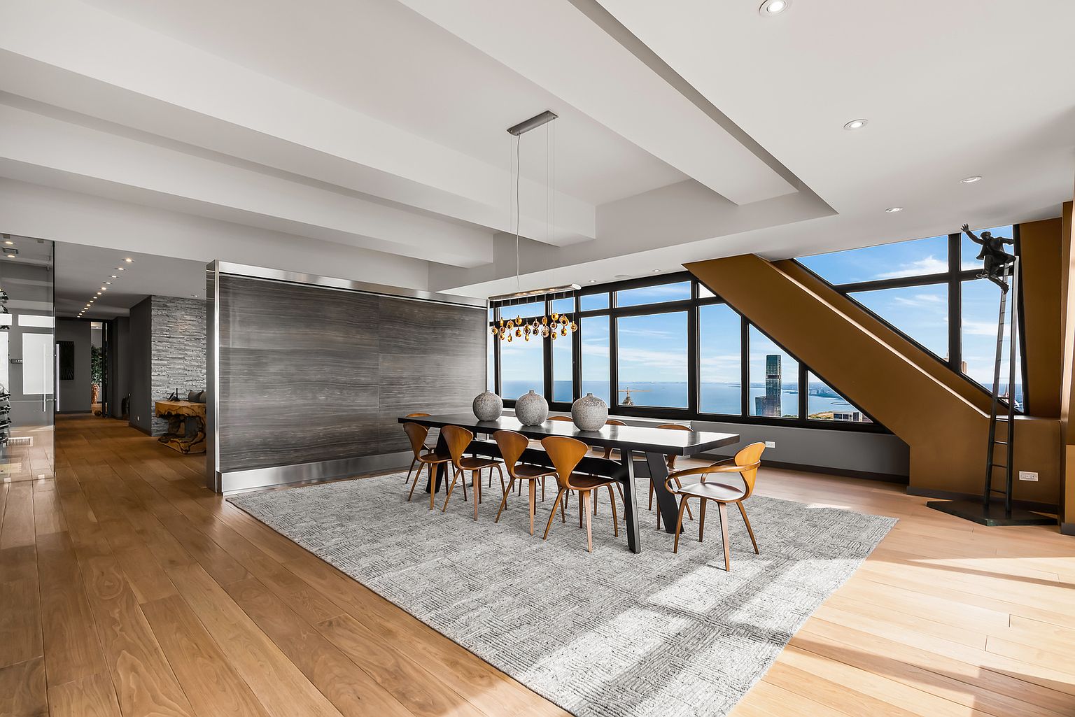 This sophisticated dining area features a long, dark-toned table surrounded by mid-century modern style chairs, all set upon a large, textured area rug. The room is defined by a striking, full-height wood-paneled wall and expansive windows that offer panoramic city and water views. A unique sculptural art piece stands near the window, adding a dramatic focal point to the open-concept space.