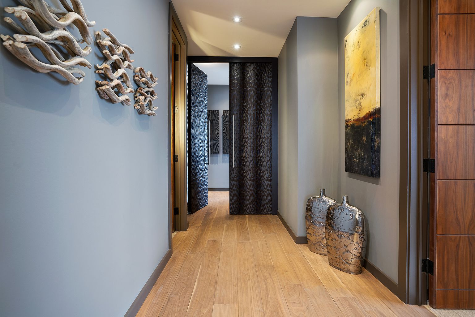 This sophisticated hallway features light wood flooring and walls painted in a muted blue-gray tone. The space is adorned with sculptural wall art and a tall, abstract painting, while two metallic vases sit on the floor near a dark wood door. A textured, dark-toned sliding door leads into an adjacent room, creating a dramatic contrast in this modern, well-lit corridor.