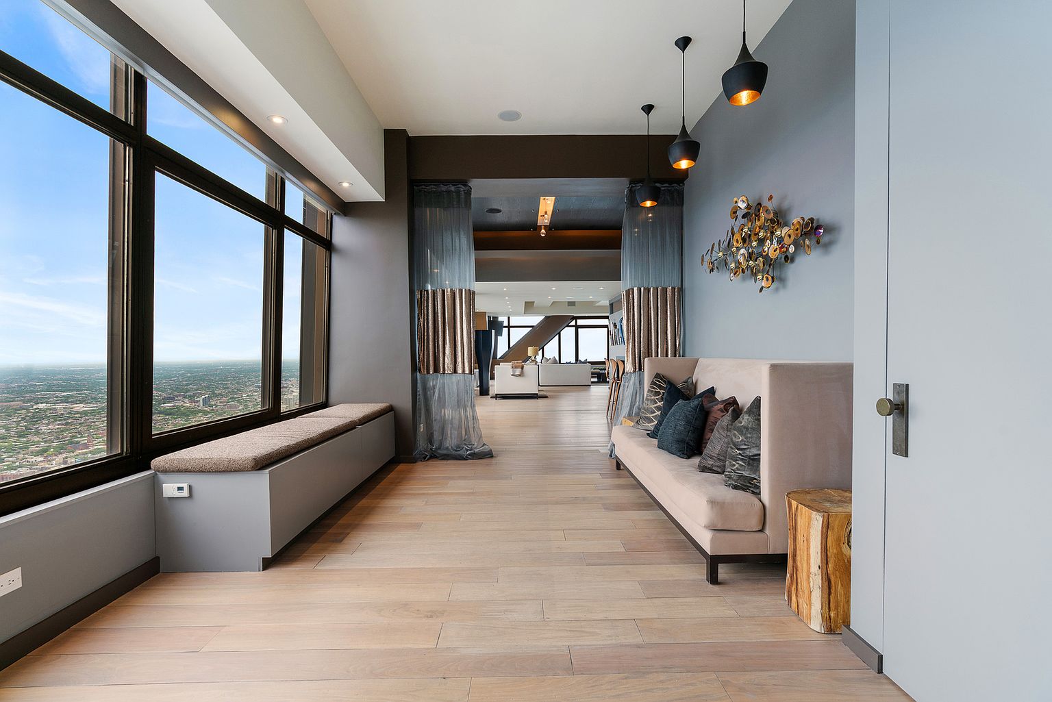 This sophisticated hallway features light wood flooring and a long, built-in window seat that offers expansive city views through large windows. A plush, neutral-toned sofa sits against a soft blue wall adorned with a decorative metallic wall sculpture, while three modern pendant lights hang above. The space leads through a draped transition area into a bright, open-concept living space, creating a sense of depth and luxury.