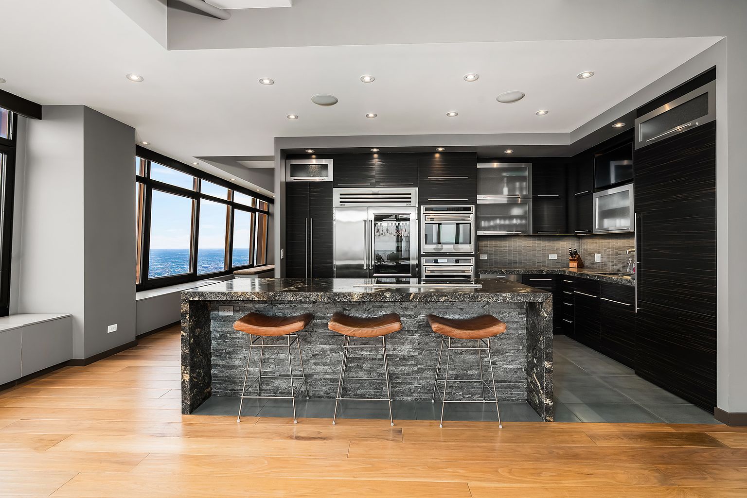This modern, high-end kitchen features a large central island with a stone waterfall edge and a textured stone base, paired with three leather bar stools. The space is defined by dark, sleek cabinetry, stainless steel appliances, and a wall of large windows that provide an expansive, elevated view of the city skyline. The combination of warm wood flooring and dark, sophisticated finishes creates a luxurious and contemporary atmosphere.