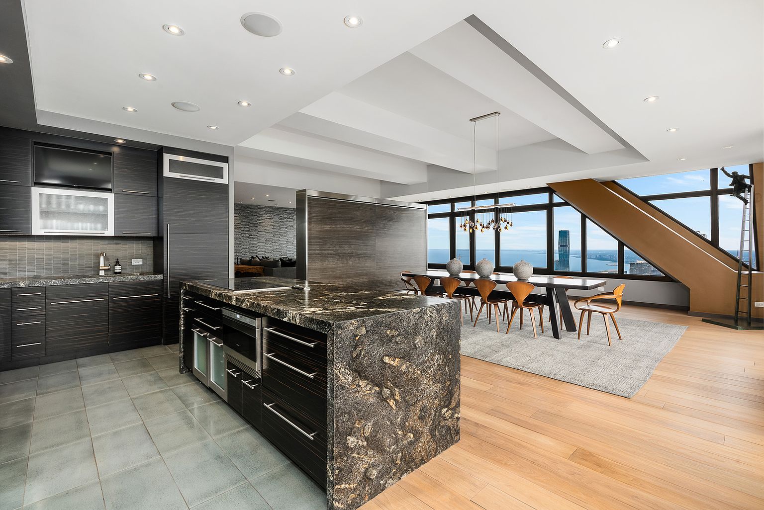 This expansive, modern open-concept kitchen and dining area features a large island with a dark, textured stone countertop and dark wood cabinetry. The dining space is positioned near large windows that offer panoramic city and water views, complemented by a unique sculptural art piece near the window frame. The room is defined by a clean, contemporary aesthetic with a mix of light wood flooring and neutral tile, creating a sophisticated and airy atmosphere.