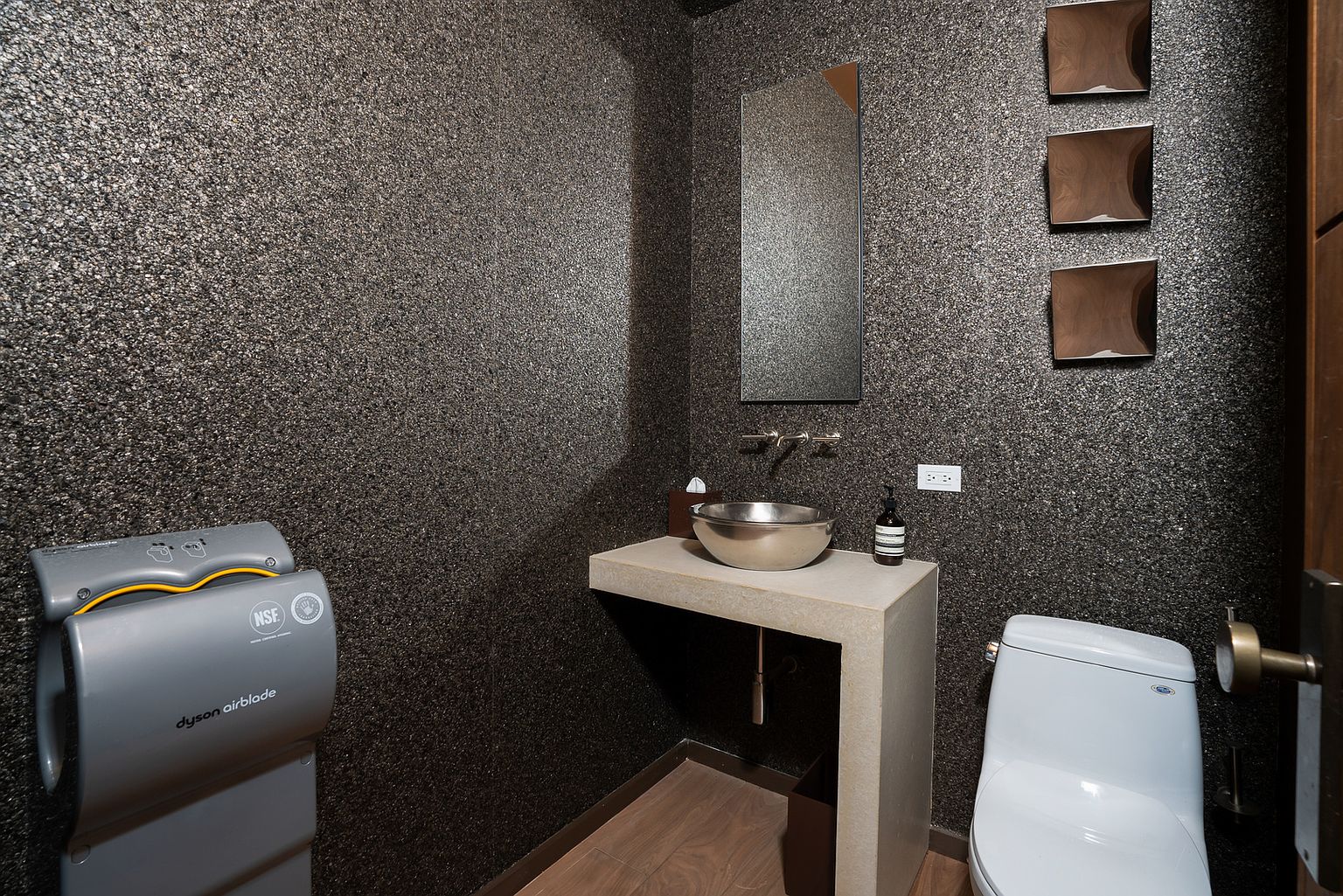 This compact powder room features a striking, dark textured wall finish that provides a dramatic backdrop for the minimalist design. A sleek, metallic vessel sink sits atop a simple, light-toned floating vanity, complemented by a wall-mounted mirror and a Dyson Airblade hand dryer. The space is finished with wood-look flooring and three square decorative wall accents, creating a modern and high-end aesthetic.