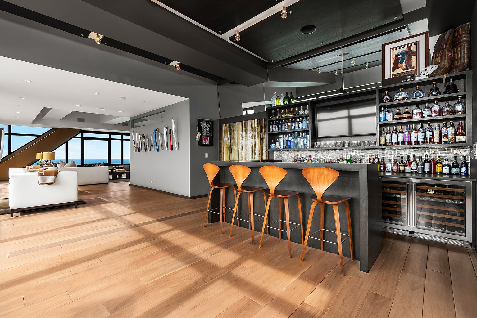 This image showcases a sophisticated home bar area featuring a dark, sleek bar counter with four modern wooden stools. The back bar is fully stocked with an extensive collection of spirits and includes a built-in wine refrigerator, while the adjacent wall displays a collection of sports memorabilia. The open-concept layout flows seamlessly into a bright living area with large windows that provide a panoramic view of the outdoors.