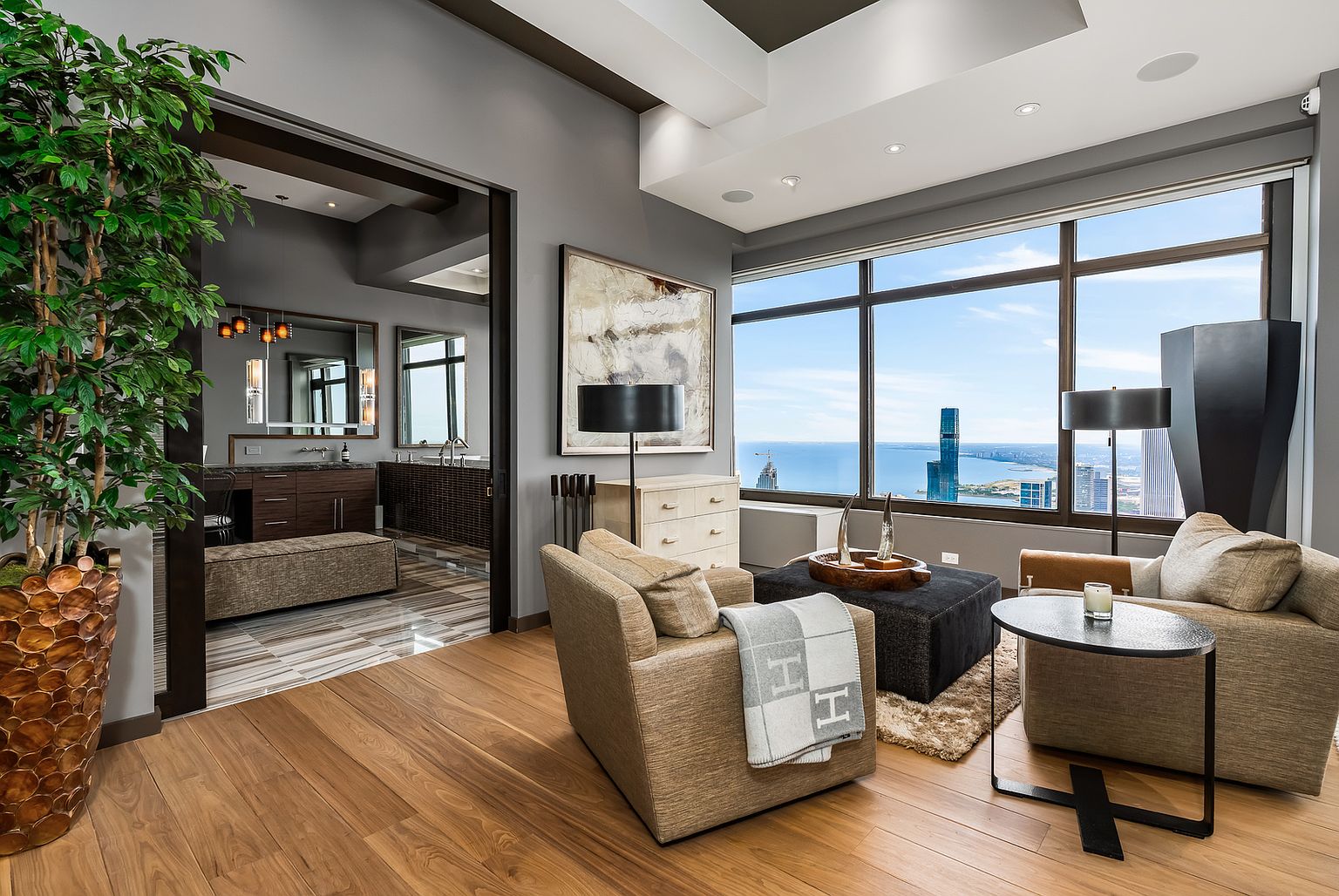 This sophisticated living area features a pair of comfortable armchairs arranged around a dark ottoman, creating an intimate seating nook. Large windows provide an expansive, elevated view of the city skyline and the water beyond, while a doorway leads into a well-appointed bathroom with dark wood cabinetry. The space is accented by a tall potted plant, a textured area rug, and a piece of abstract wall art, conveying a luxurious and modern urban aesthetic.