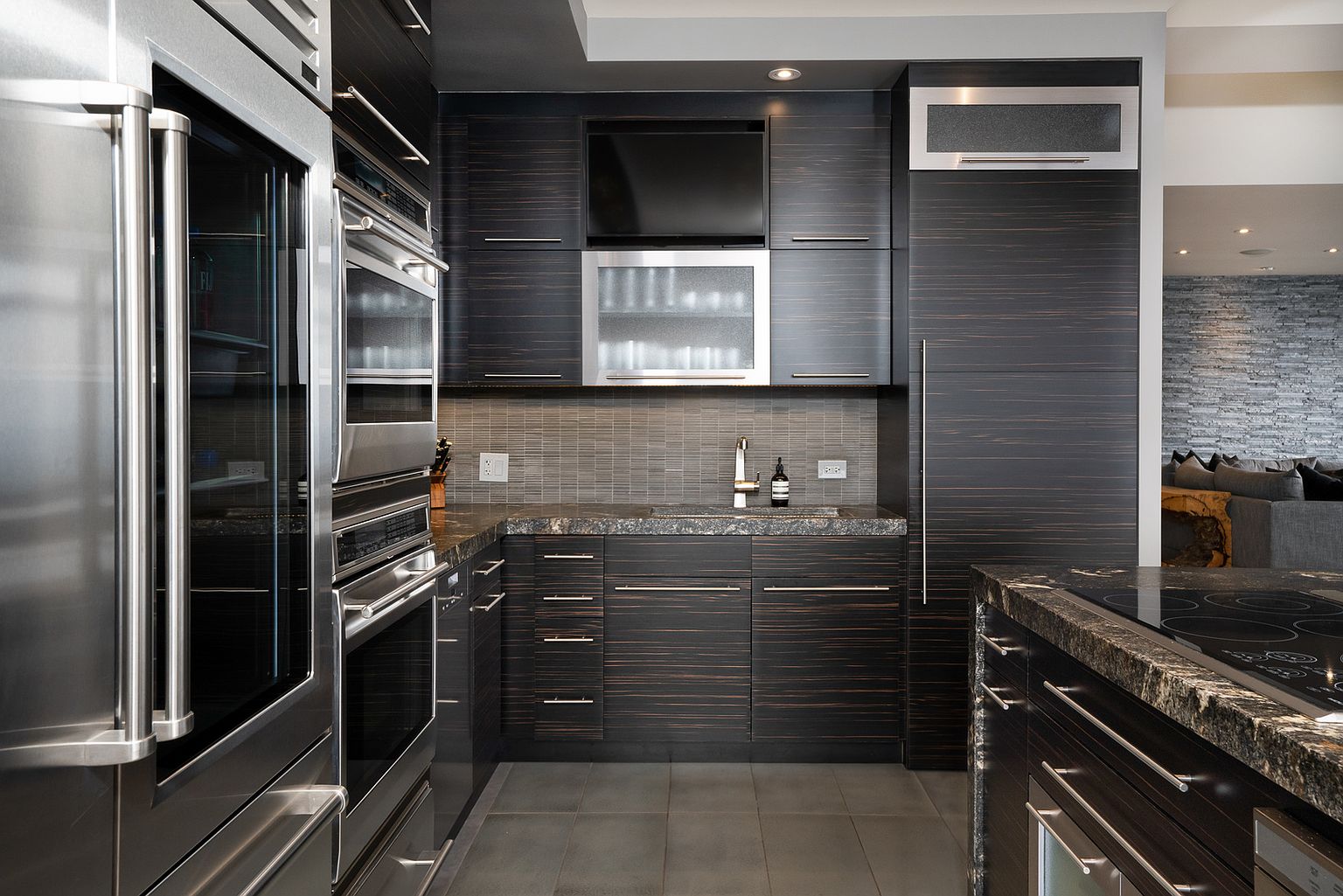 This sleek, contemporary kitchen features dark, horizontal-grain cabinetry paired with dark stone countertops and a tiled backsplash. The space is equipped with high-end stainless steel appliances, including a built-in wall oven and a large refrigerator, creating a sophisticated and functional culinary environment. The perspective captures the efficient galley-style layout, highlighting the seamless integration of modern fixtures and professional-grade equipment.