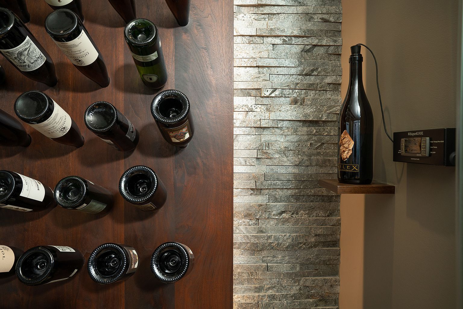 This image captures a sophisticated wine storage area, featuring a dark wood wall with individual bottle cradles arranged in a grid pattern. Adjacent to the wine rack is a textured, stacked-stone wall that provides a rustic contrast, with a single bottle displayed on a small wooden shelf. A digital temperature control unit is mounted on the wall, emphasizing the climate-controlled nature of this refined space.