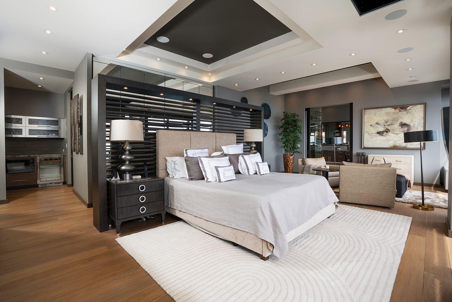 This sophisticated primary bedroom features a central bed positioned against a dark, slatted wood room divider that separates the sleeping area from a small kitchenette. The room is decorated in a neutral palette with soft textures, including a large patterned area rug and plush bedding, while recessed lighting and a tray ceiling add architectural depth. To the side, a cozy seating area with a sofa and a tall floor lamp creates a comfortable retreat, complemented by a large piece of abstract wall art.