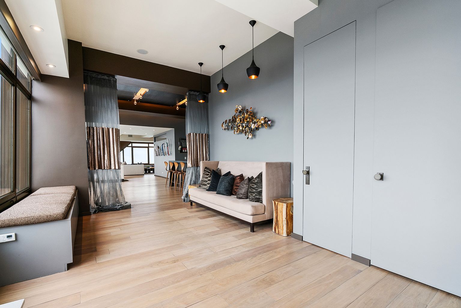 This sophisticated living area features light wood flooring and a neutral-toned sofa adorned with patterned throw pillows. A unique metallic wall sculpture serves as a focal point above the sofa, while three modern pendant lights hang from the ceiling. The space flows seamlessly into an adjacent room through an opening framed by elegant, sheer curtains, creating an open and airy atmosphere.