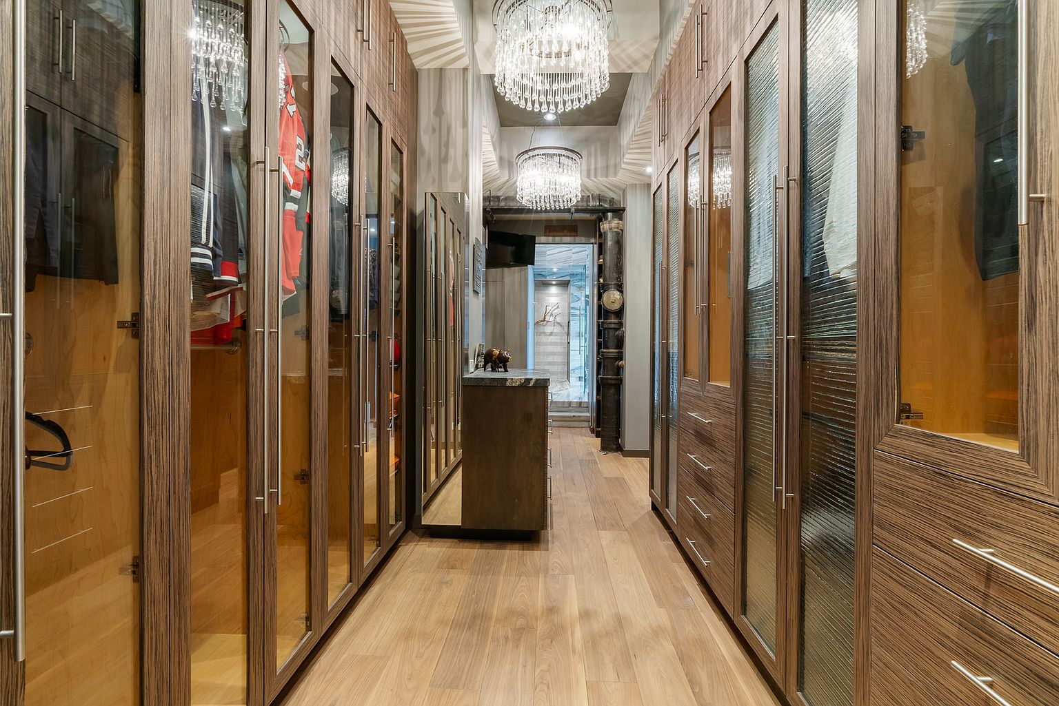 This luxurious walk-in closet features extensive custom cabinetry with glass-front doors and integrated lighting, creating a sophisticated and organized storage space. The room is anchored by a central island with a stone countertop and is illuminated by two ornate crystal chandeliers. The perspective looks down the center aisle, highlighting the rich wood-toned finishes and the seamless transition into the adjacent room.