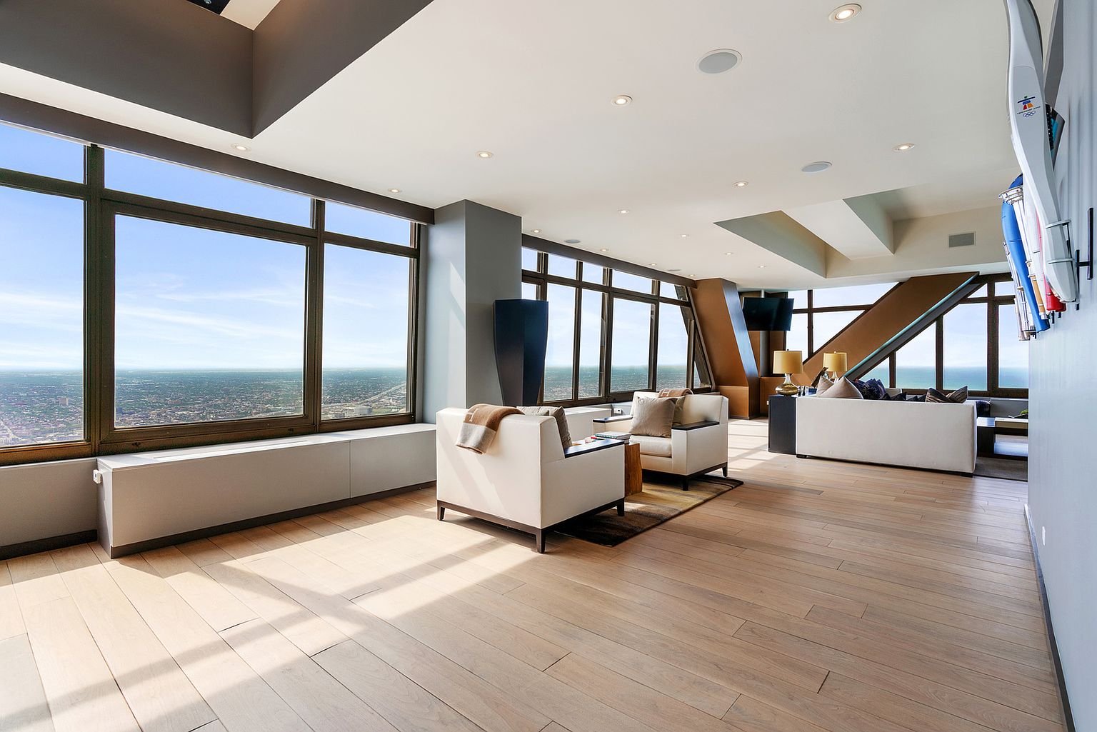 This expansive, high-rise living area features light wood flooring and a minimalist aesthetic, anchored by two cream-colored armchairs and a sofa set. Large windows wrap around the perimeter, offering panoramic views of the city skyline and the horizon. The open-concept layout is illuminated by recessed lighting and natural light, creating a bright, airy, and sophisticated atmosphere.