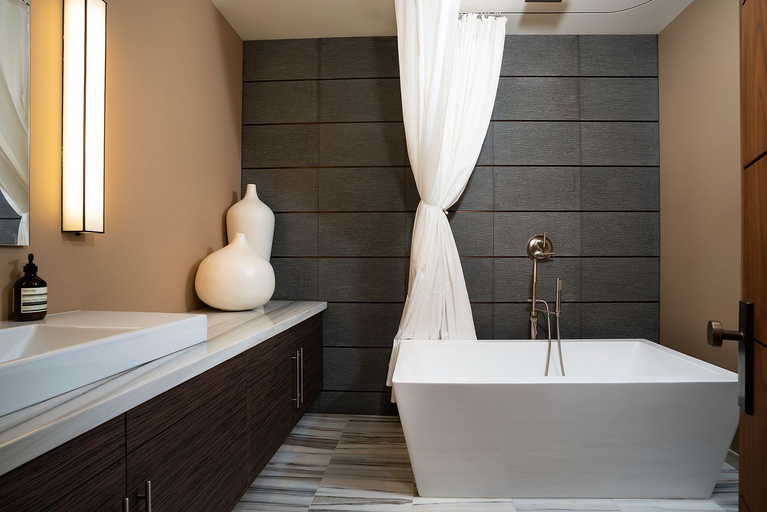 This modern bathroom features a sleek, freestanding white soaking tub positioned against a textured, dark-toned tiled wall. To the left, a long vanity with a stone countertop and dark wood cabinetry provides ample storage, complemented by a minimalist light fixture. The space is finished with patterned stone flooring, creating a sophisticated and tranquil spa-like atmosphere.