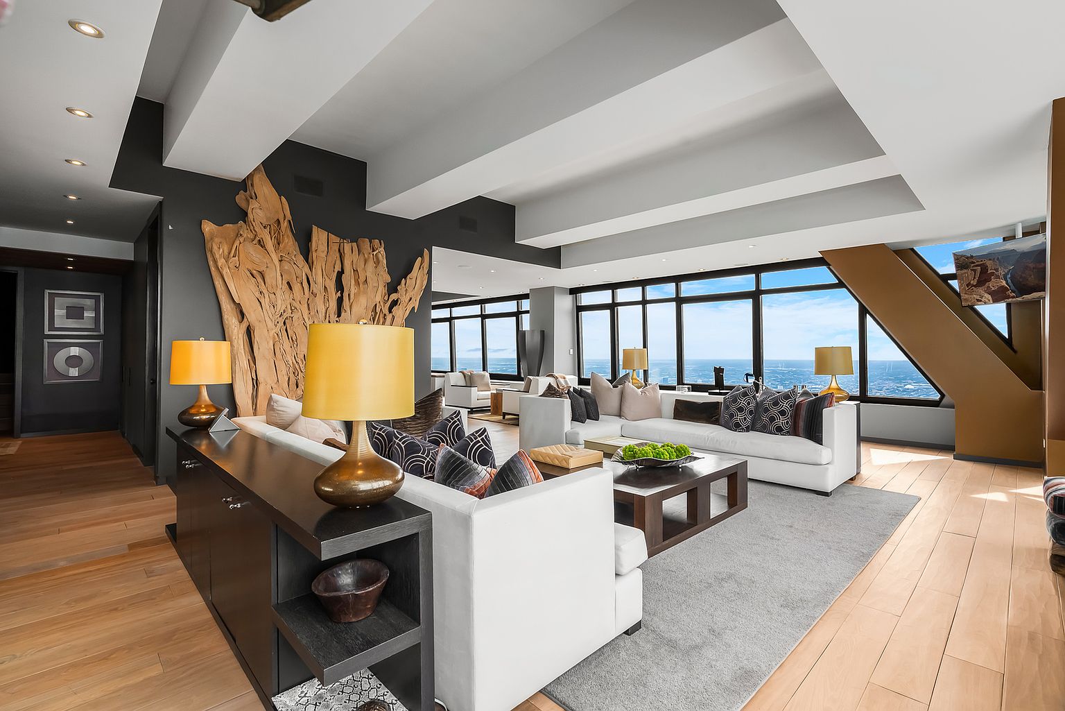 This expansive, modern living room features a sophisticated open-concept layout with light wood flooring and a striking, large-scale natural wood sculpture mounted on a dark accent wall. The space is anchored by two white sectional sofas arranged around a dark wood coffee table, all set atop a large gray area rug. Large windows provide panoramic views of the city and horizon, while recessed lighting and stylish table lamps create a warm, inviting atmosphere.