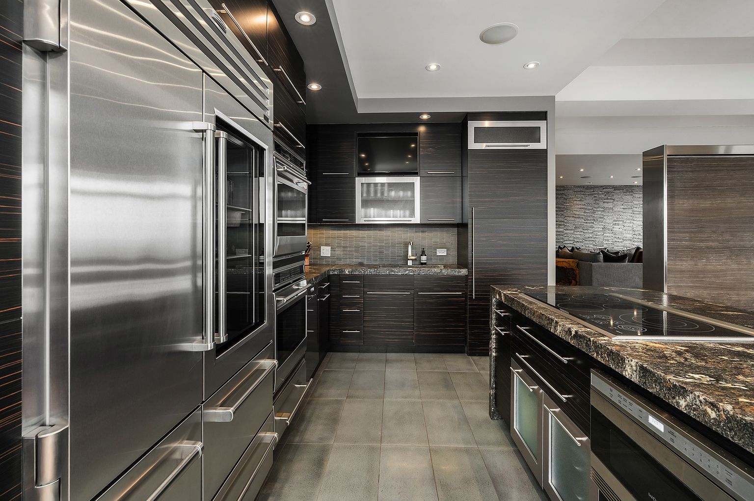 This sleek, modern kitchen features dark, horizontal-grain cabinetry paired with dark stone countertops and stainless steel appliances. The galley-style layout includes a large island with an integrated cooktop and a built-in oven, while the floor is finished with large, neutral-toned tiles. The perspective looks down the narrow corridor toward the sink area, showcasing a sophisticated, high-end aesthetic.