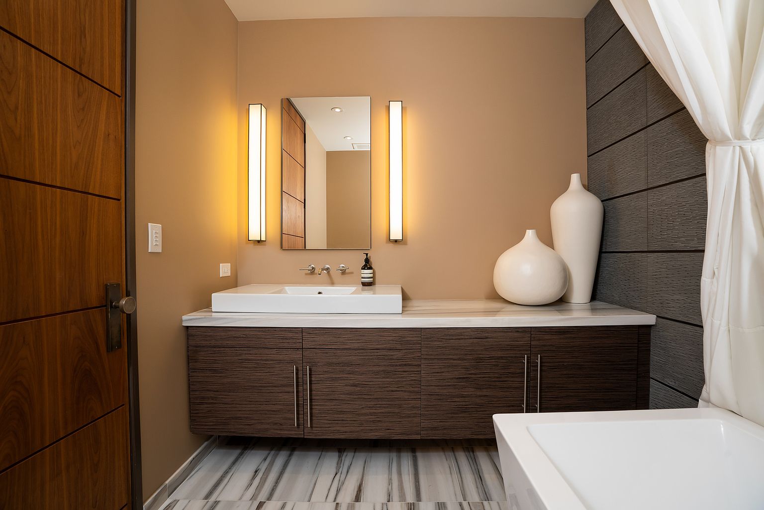 This modern bathroom features a floating vanity with dark, textured cabinetry topped by a white stone counter and a rectangular vessel sink. The space is accented by warm, vertical wall-mounted lighting flanking a central mirror, while two large, minimalist white vases add a sculptural element to the counter. The room is finished with a striking striped stone floor and a dark, horizontally paneled accent wall, creating a sophisticated and contemporary atmosphere.