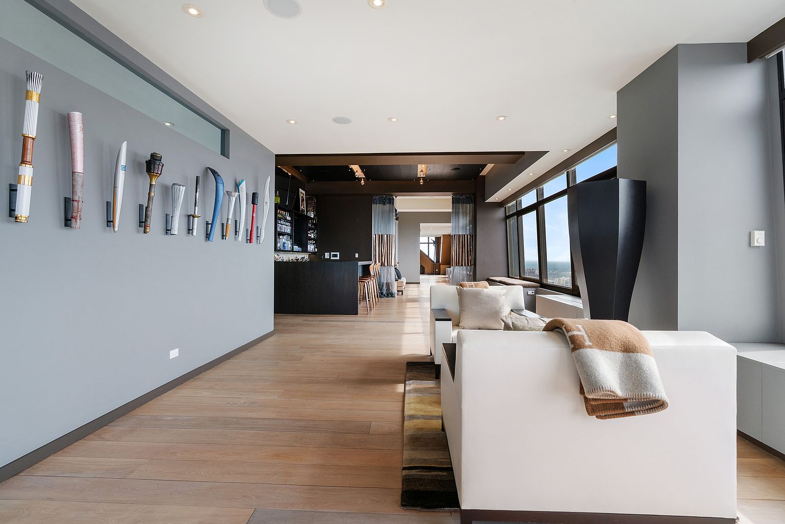 This sophisticated living area features a unique gallery wall displaying a collection of Olympic torches, creating a distinct focal point against the neutral gray wall. The space flows seamlessly into a bar area, with light wood flooring throughout and a white sofa accented by a patterned throw blanket. Large windows along the side provide expansive views and fill the room with natural light, contributing to an open and airy atmosphere.