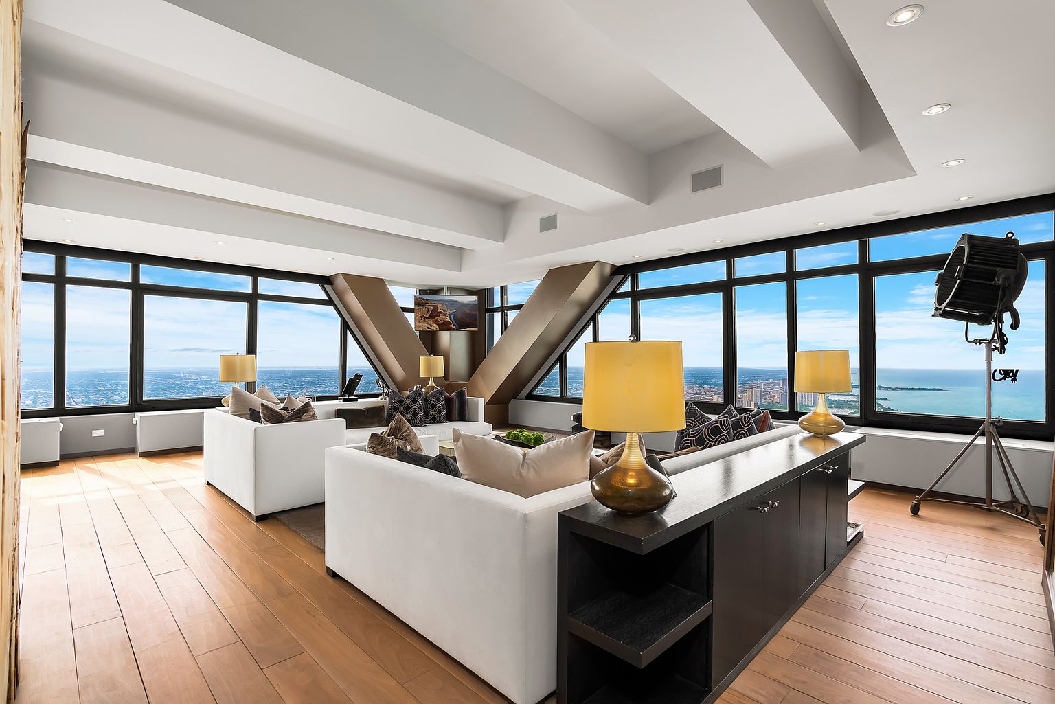 This expansive, high-rise living room features a contemporary open-concept layout with light wood flooring and neutral-toned sofas. The space is defined by its dramatic, angled floor-to-ceiling windows that provide panoramic views of the city skyline and the lake. A unique industrial-style studio spotlight on a tripod adds a bold, artistic touch to the sophisticated, modern interior.