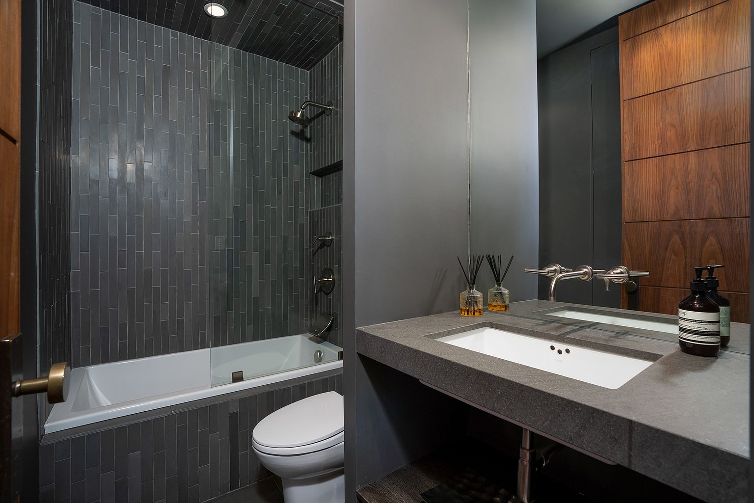 This modern bathroom features a sleek, dark aesthetic with vertical charcoal-toned tiles lining the shower and tub area. A floating vanity with a thick, dark stone countertop and a wall-mounted faucet provides a minimalist look, complemented by a wood-paneled wall and a large mirror. The space is efficiently designed, incorporating a toilet and a glass-enclosed shower-tub combination in a compact, sophisticated layout.