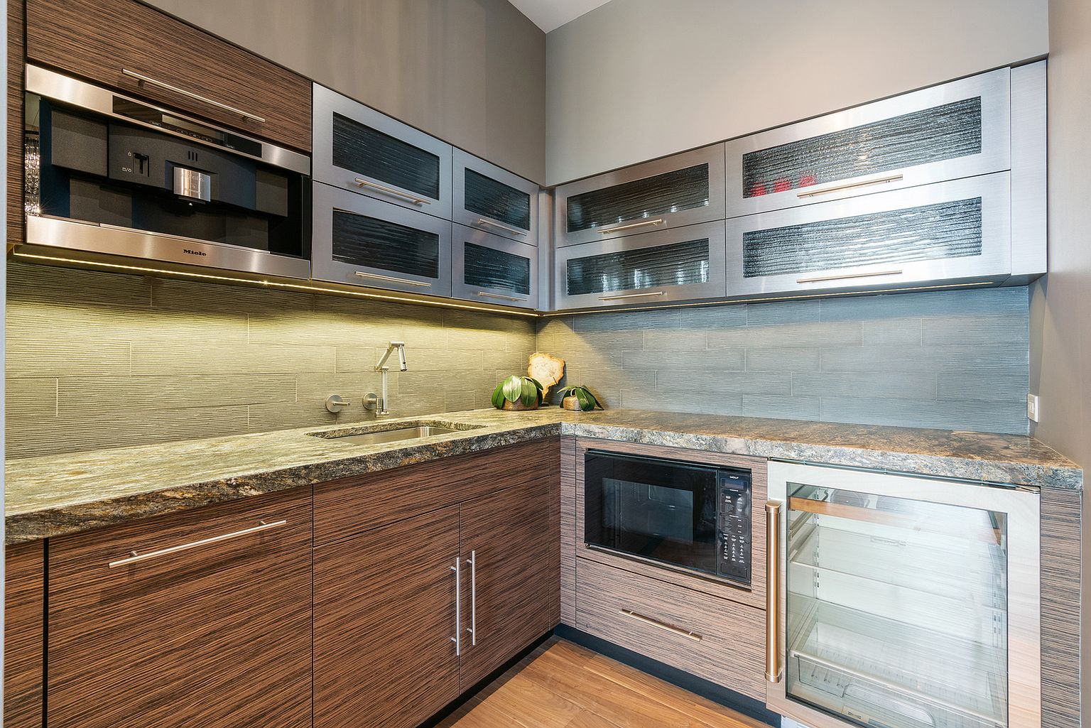 This modern kitchen area features sleek, horizontal-grain wood cabinetry paired with metallic-framed glass-front upper cabinets. The space includes a built-in coffee machine, a microwave, and a glass-door beverage refrigerator, all set against a textured tile backsplash and stone countertops. The perspective is a wide-angle shot capturing the L-shaped layout, emphasizing the high-end appliances and contemporary design aesthetic.