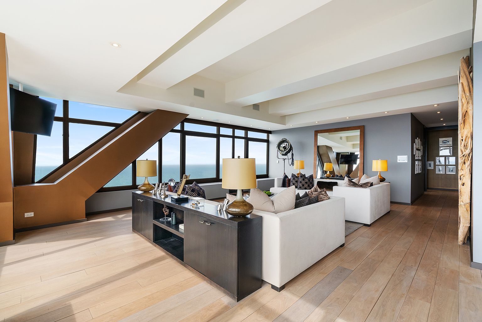 This expansive, modern living room features light wood flooring and a neutral color palette, anchored by two white sofas arranged around a dark console table. Large windows provide panoramic ocean views, while a unique architectural staircase structure adds a dramatic geometric element to the space. The room is accented with warm lighting from table lamps and a large mirror, creating a sophisticated and airy atmosphere.