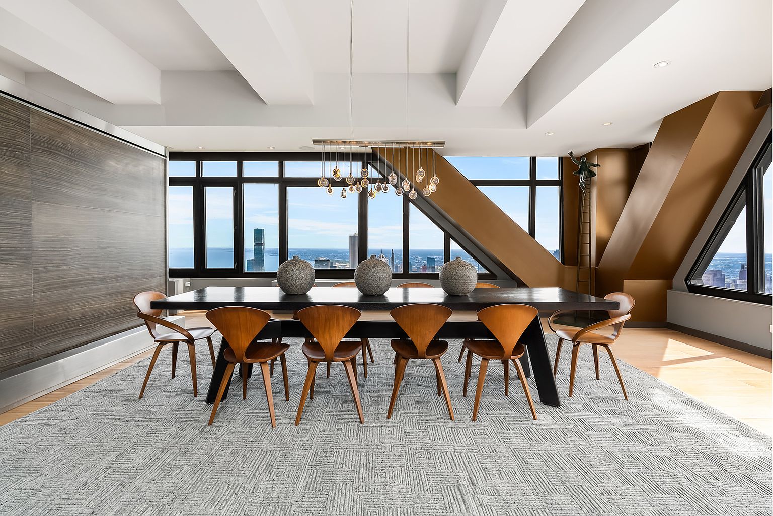 This sophisticated dining room features a long, dark-toned table surrounded by eight mid-century modern style wooden chairs, all set upon a large, textured gray area rug. The space is defined by expansive windows that offer panoramic city views and a unique, angular architectural wall finished in a metallic bronze tone. A contemporary multi-pendant light fixture hangs above the table, while the ceiling is accented with structural beams, creating a dramatic and upscale atmosphere.